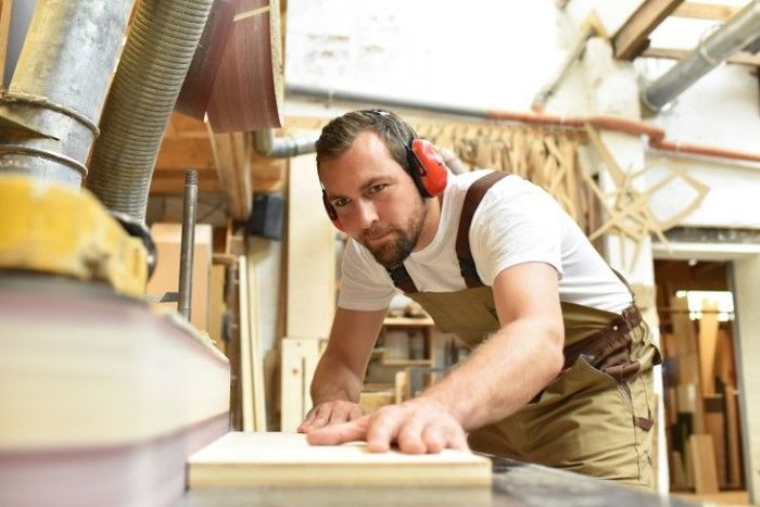 Carpenter sanding wood, wearing safety earmuffs in a workshop.