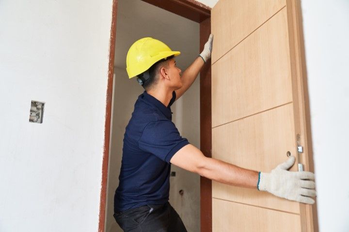 A worker in a yellow hard hat and gloves installing a wooden door in a doorway.