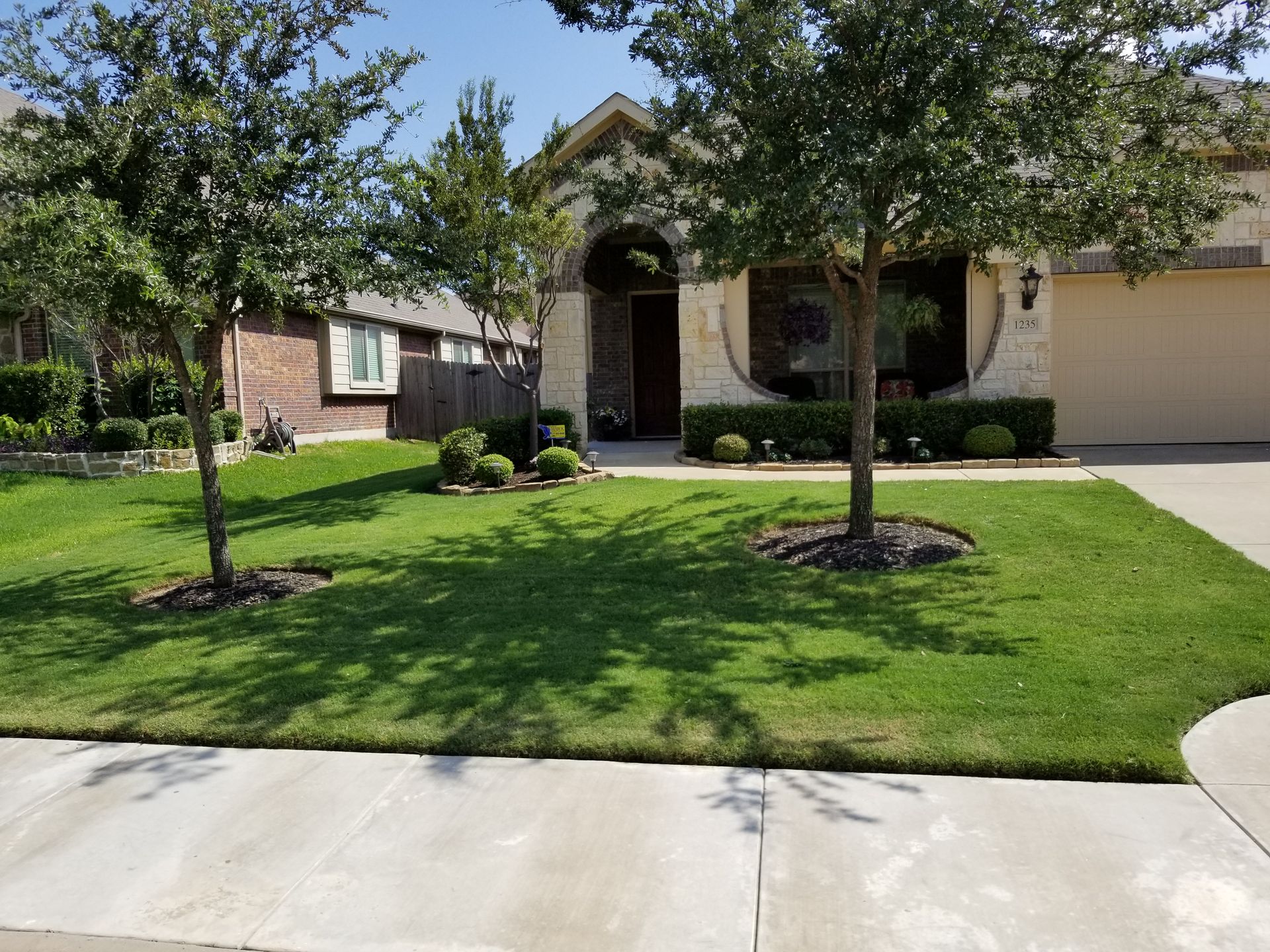 Suburban house with arched entry, two trees, and a neatly mowed front lawn by the sidewalk