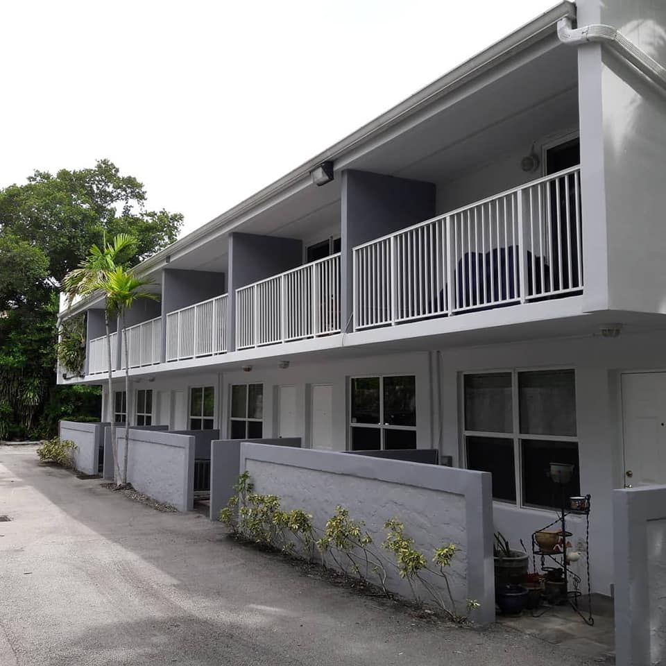 Two-story gray apartment building with white balconies and small front yards, palm tree in the background.