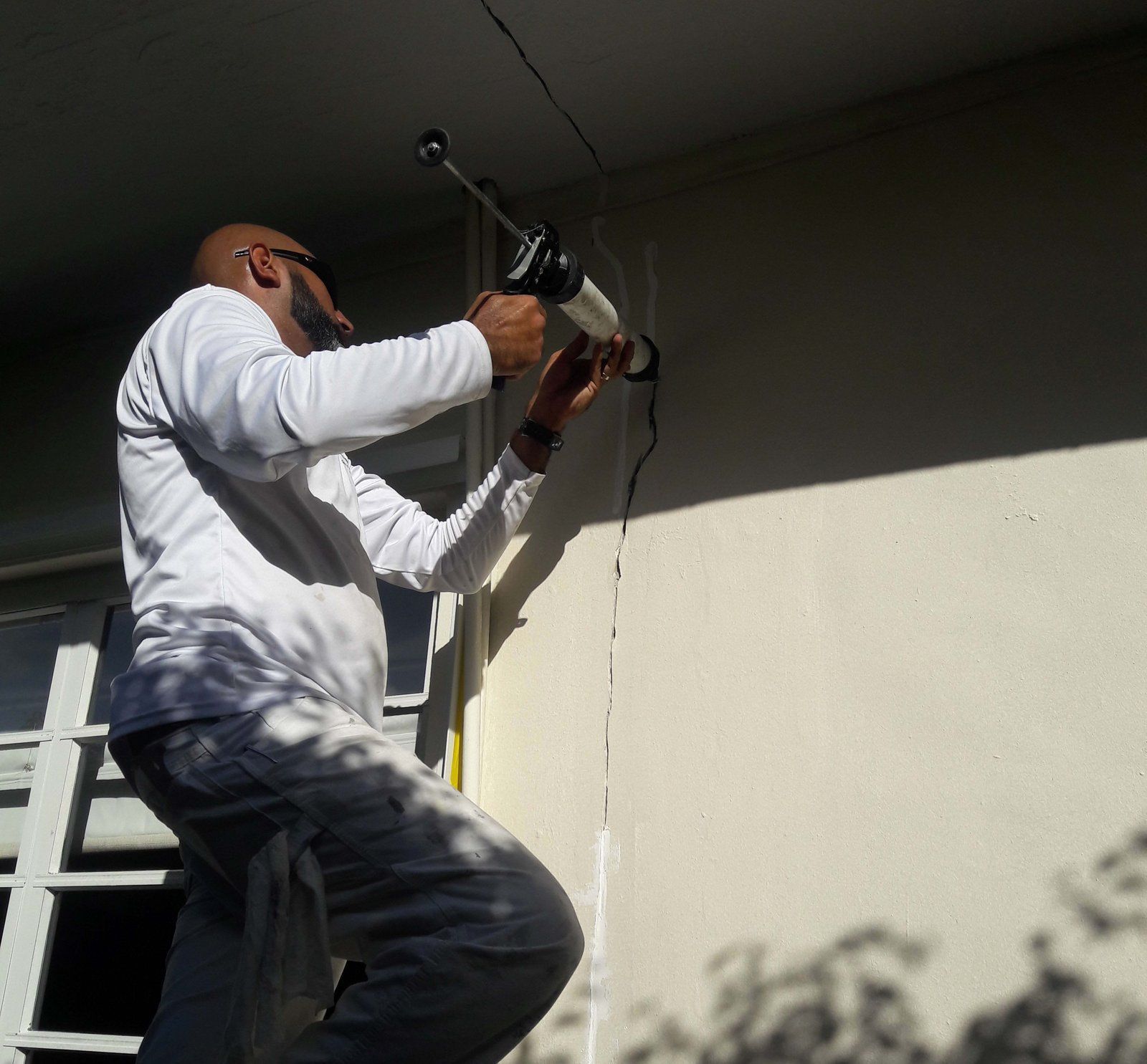 Man on ladder caulking a wall joint, holding a caulk gun.