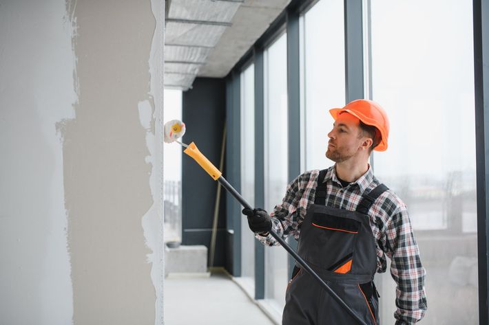 Construction worker in orange hard hat paints a white wall with a roller.