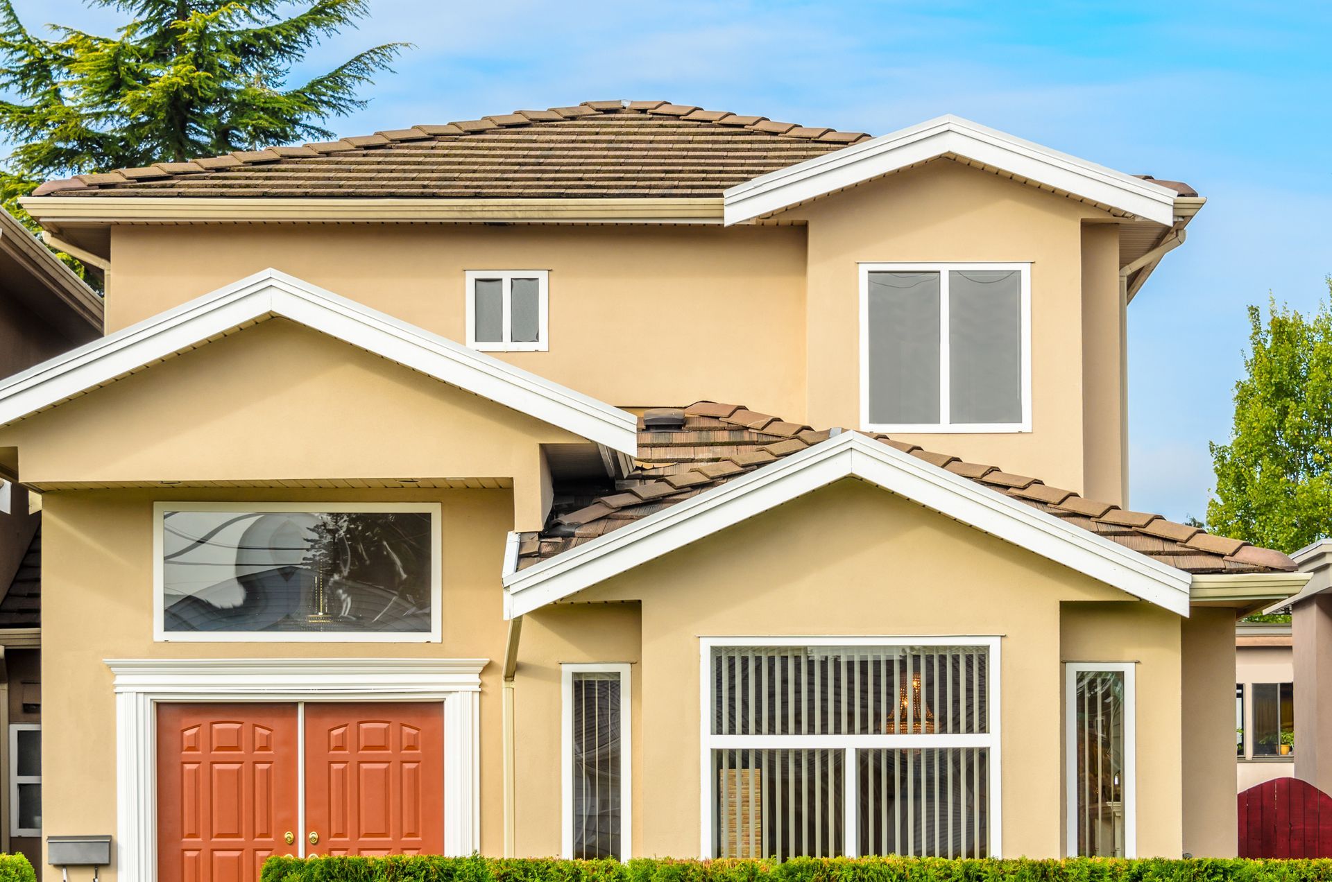 Beige two-story house with white trim, brown roof, and orange front doors against a blue sky.