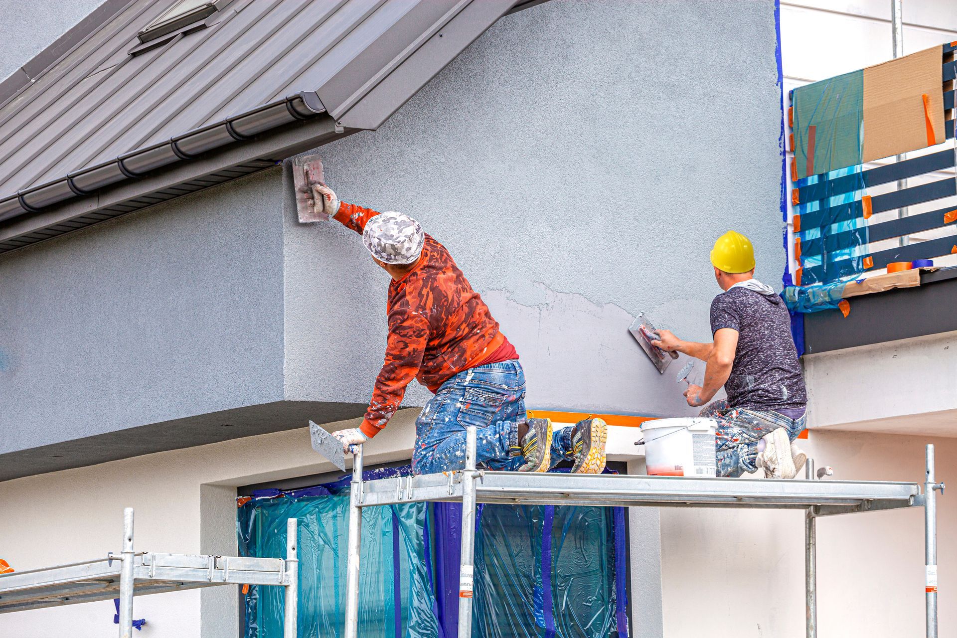 Two workers applying stucco to the exterior wall of a house; one on a scaffold.