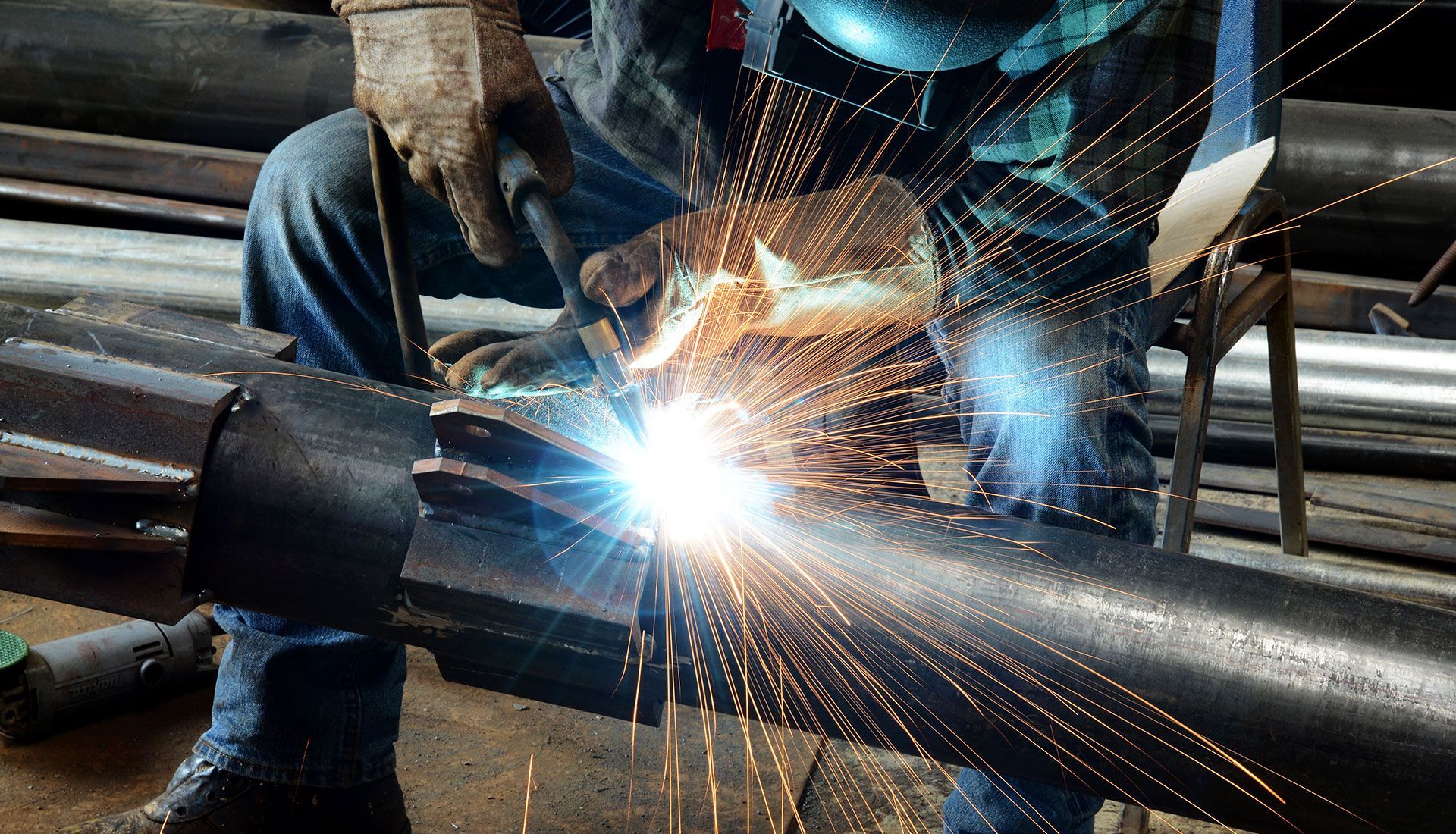 Welder working on metal pipe, sparks flying, wearing protective gear.