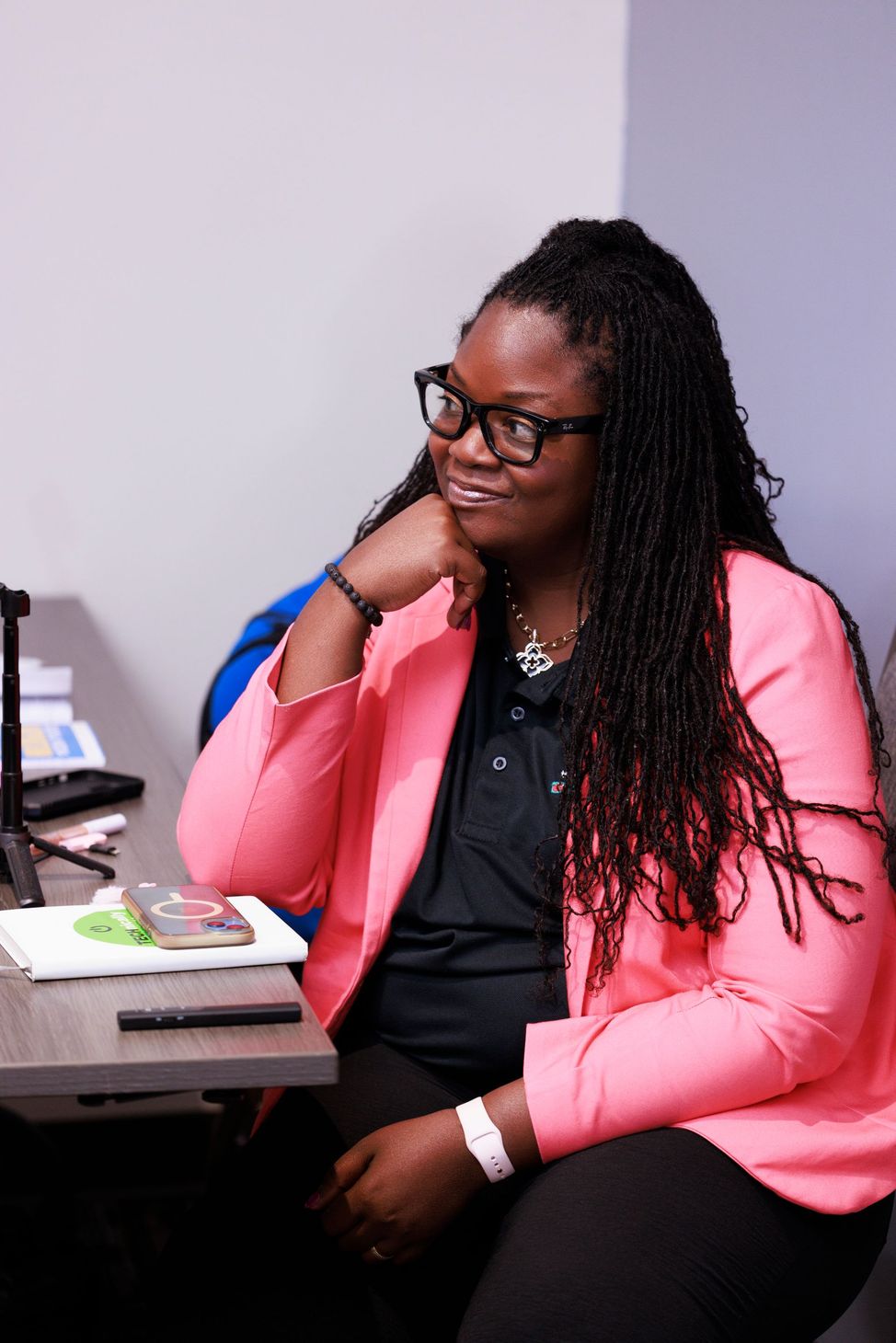 Woman in pink blazer sitting at a desk, resting her chin on her hand and looking to the side.