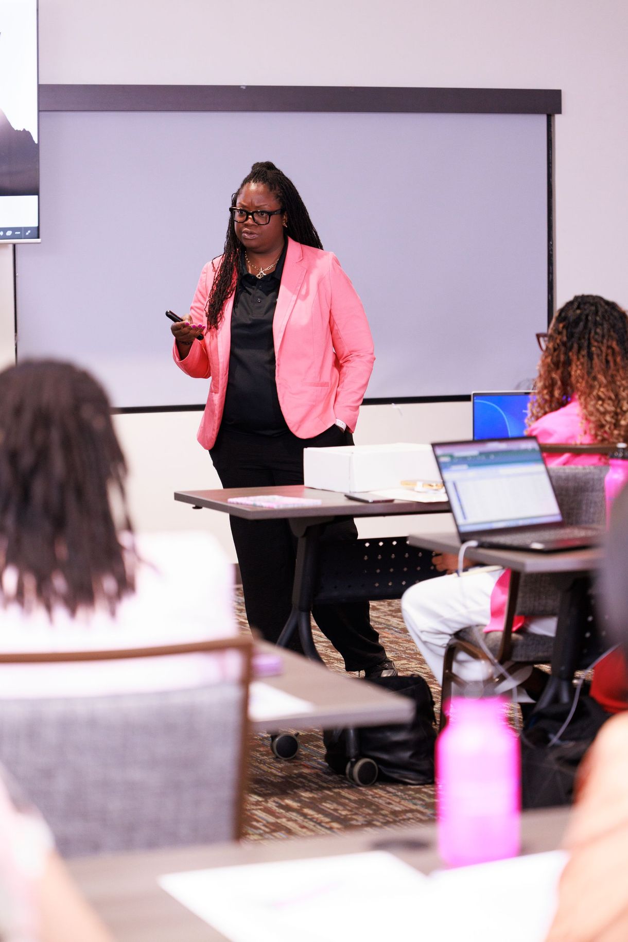Presenter in pink blazer speaking to seated audience in a classroom with laptops