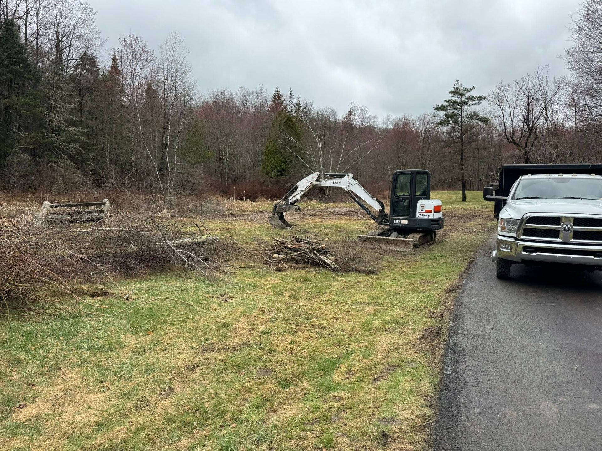 Small excavator digging soil beside a white truck on a muddy roadside under gray skies