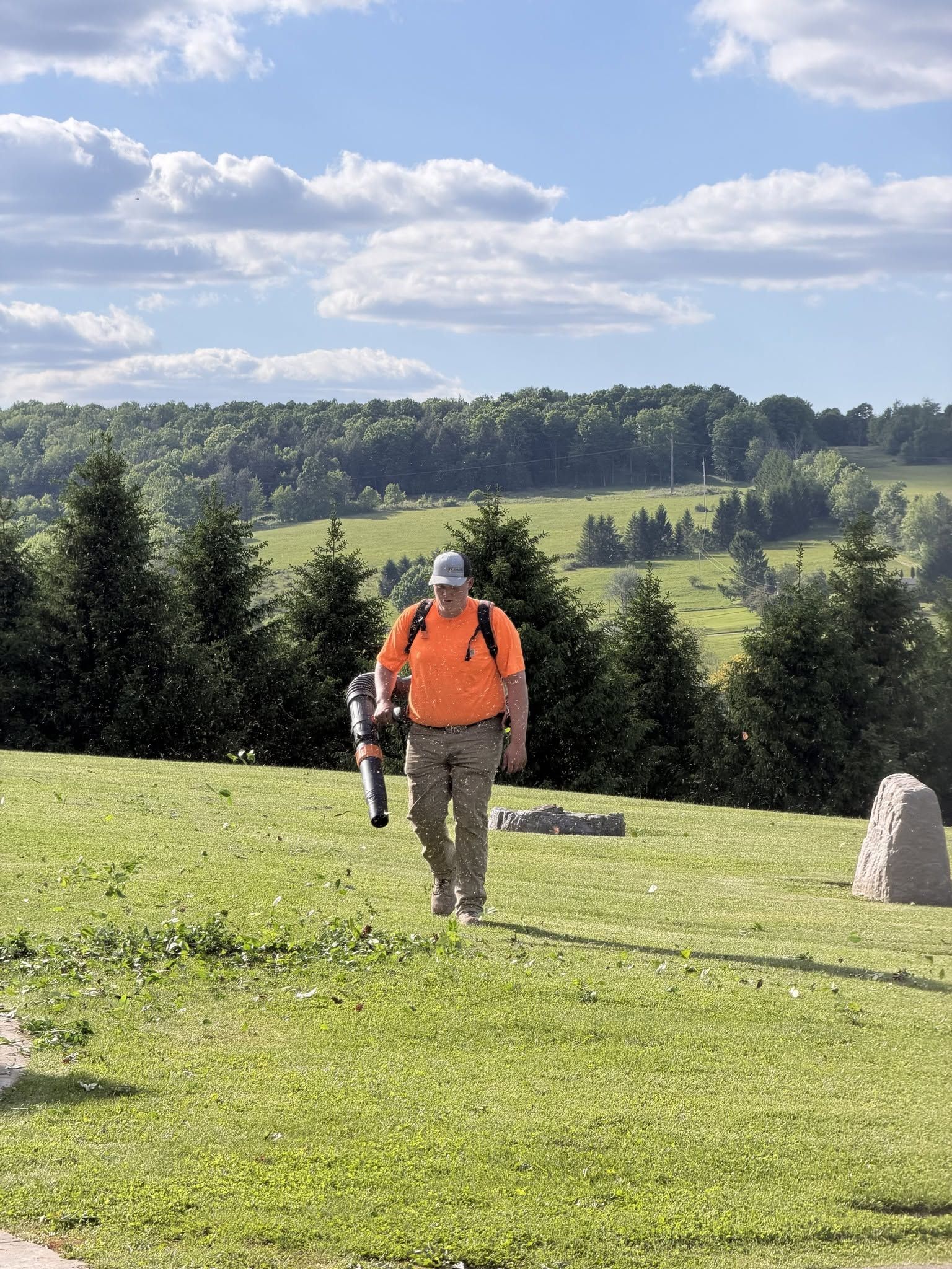 Two hikers in bright orange vests walking across a grassy hill with trees and cloudy sky in the background