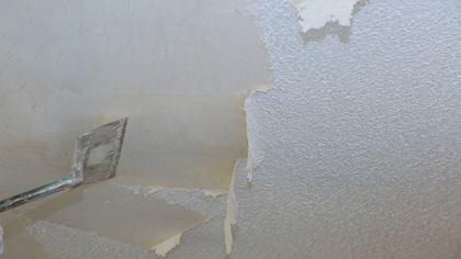 Scraper removing peeling textured ceiling paint, revealing bare drywall beneath.