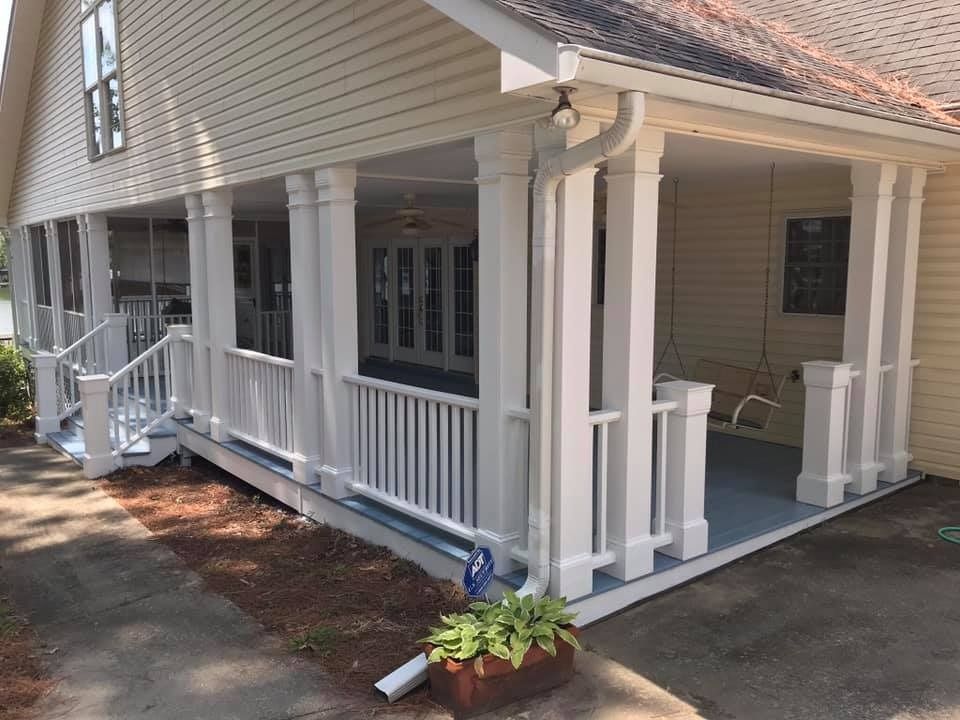 Covered porch with white columns, railings, and blue floor. Home exterior with greenery.
