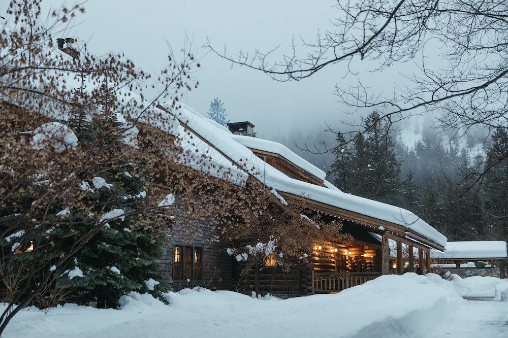 Snow-covered lodge with warm interior lights in a winter forest setting.