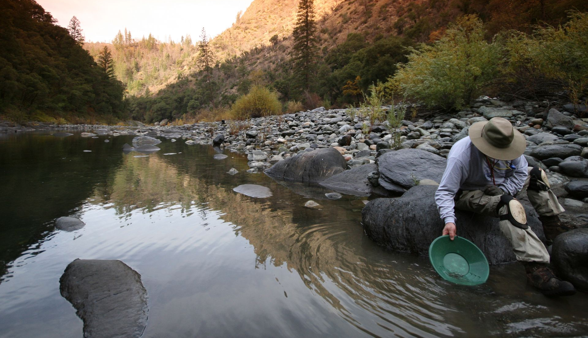 Person panning for gold beside a river in a rocky mountain landscape