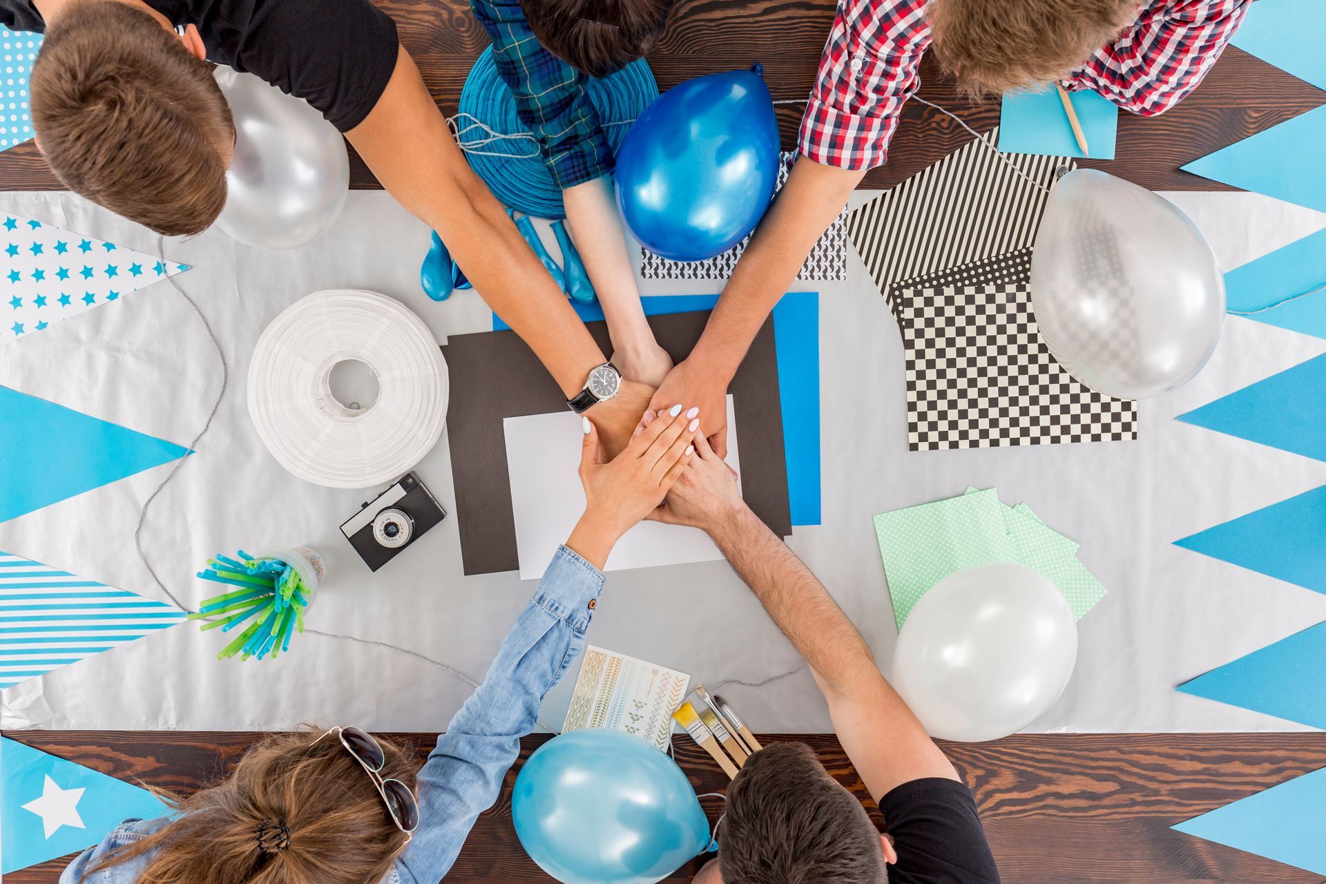 Team hands stacked over a craft table with colorful supplies and balloons, viewed from above