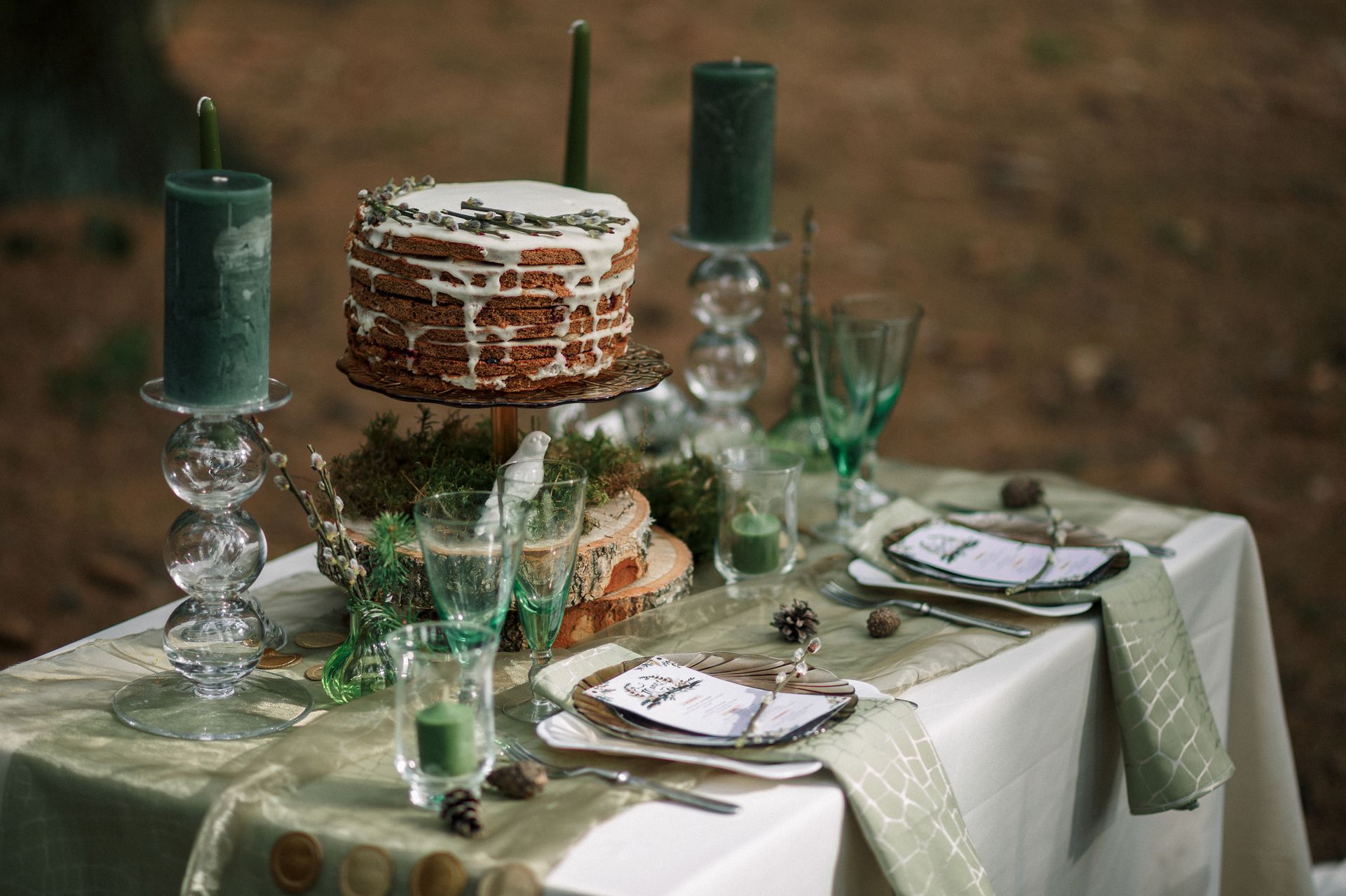 Outdoor table setting with tiered cake, green candles, and white-green place settings on a rustic tablecloth