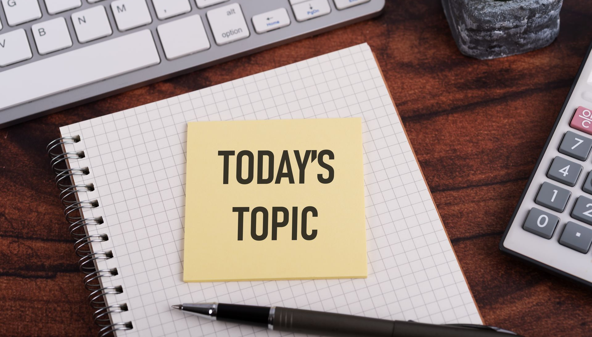 Desk with sticky note reading “TODAY’S TOPIC,” notebook, pen, keyboard, and calculator on a wooden surface.