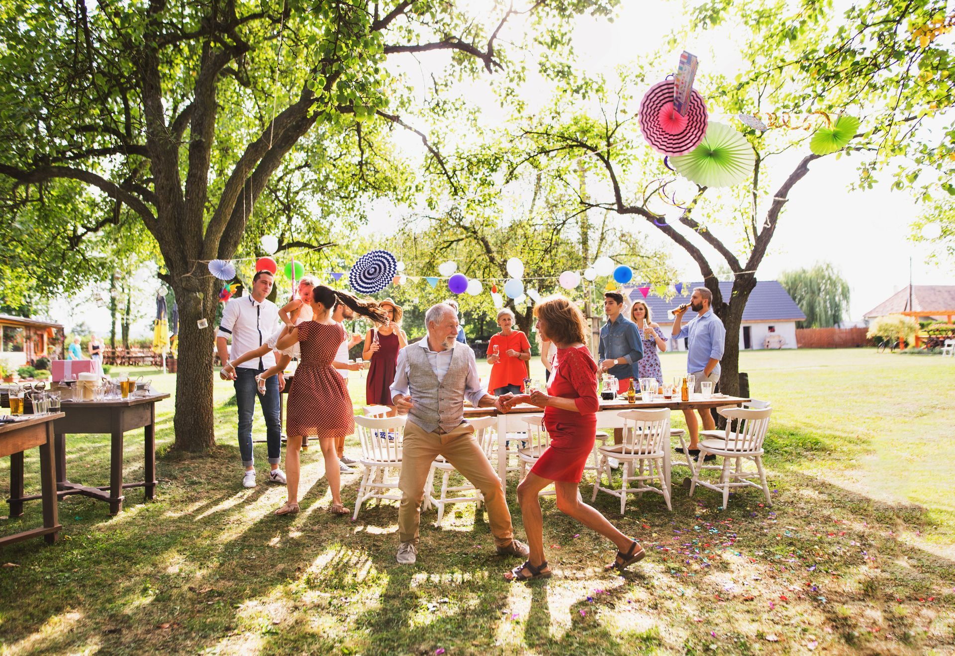 People mingling at an outdoor garden party under trees with tables, chairs, and colorful decorations