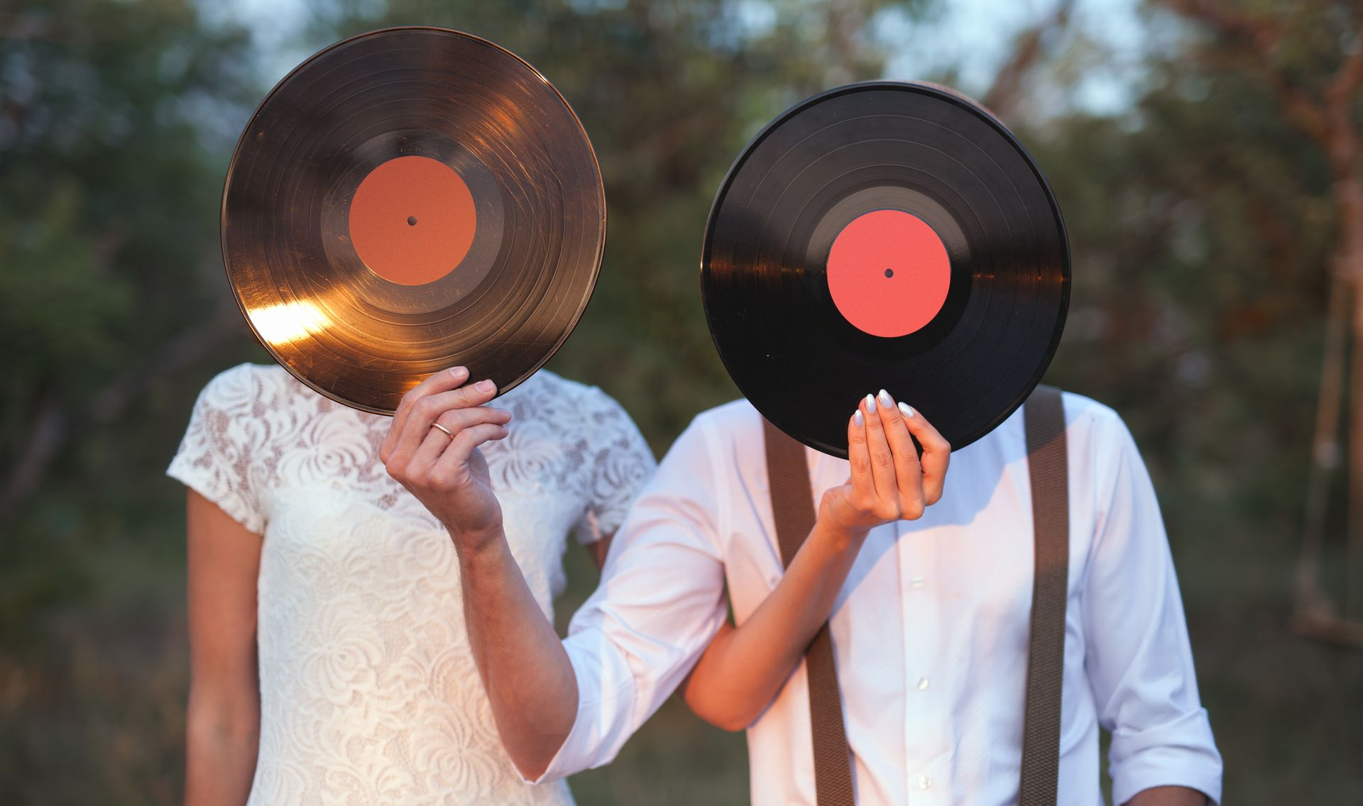 Two people holding vinyl records over their faces, standing outdoors in soft evening light.