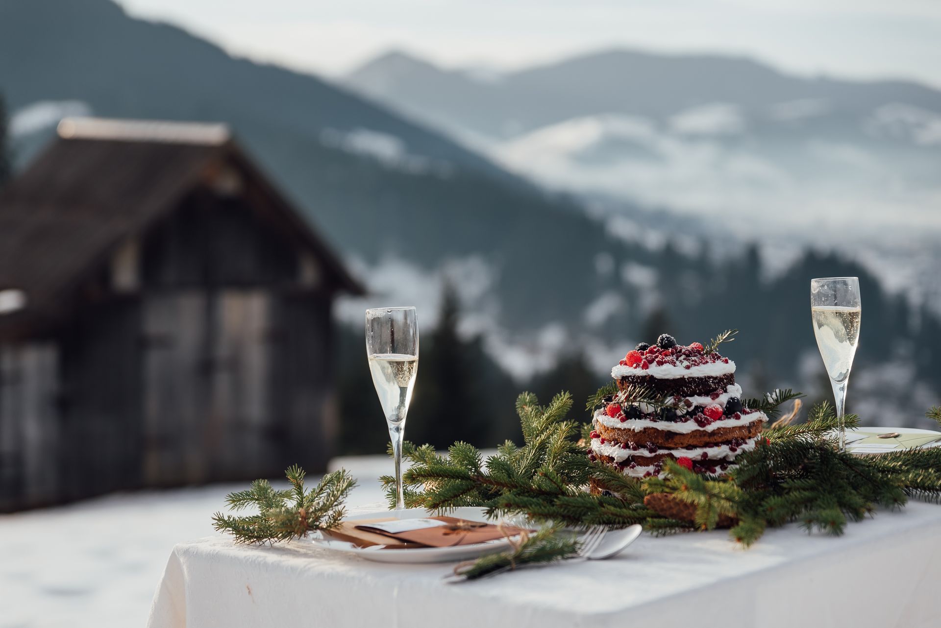Festive dessert table outdoors with cake, champagne flutes, and evergreen branches in snowy mountains