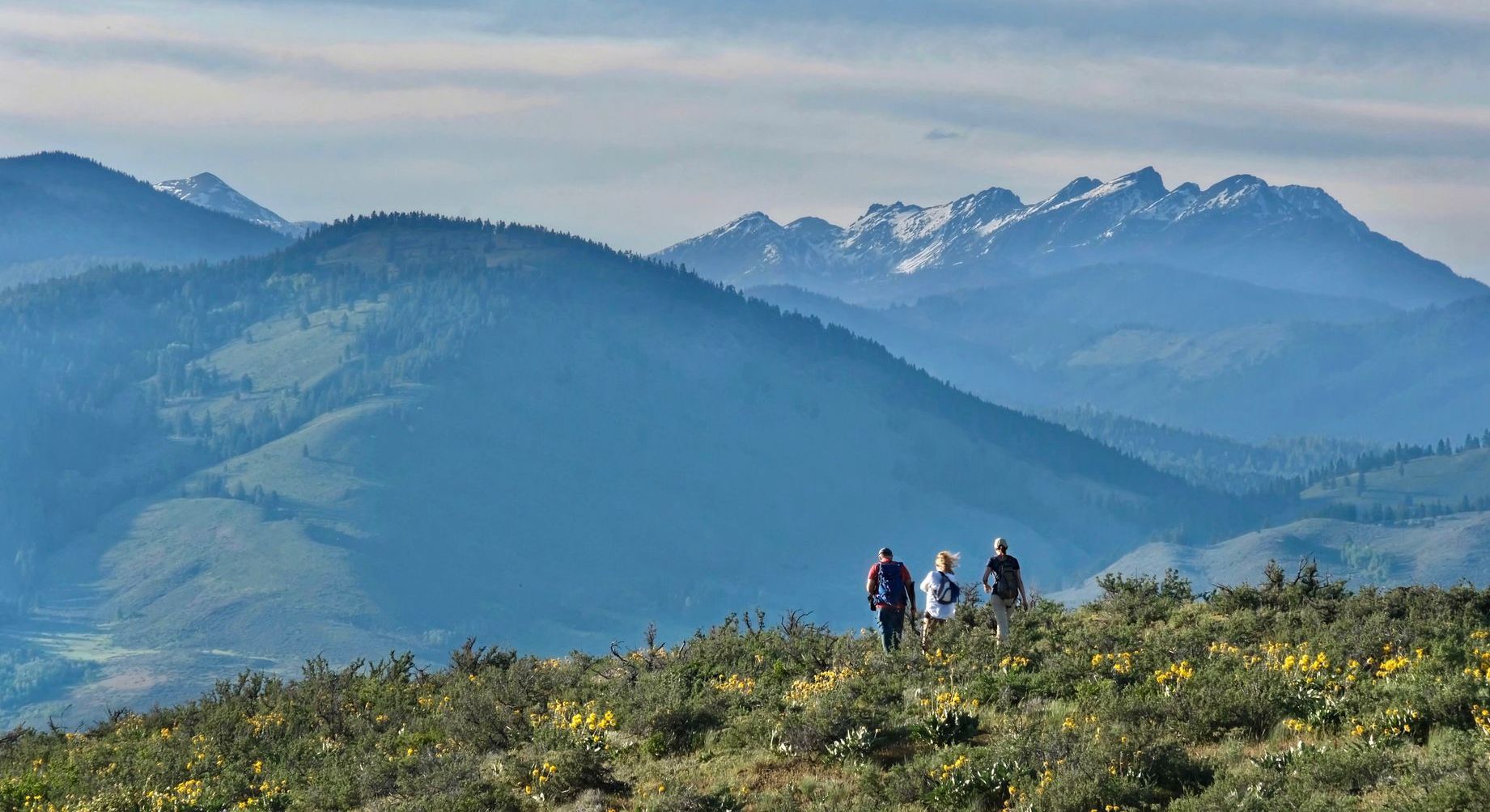 Three people are walking on top of a hill with mountains in the background.