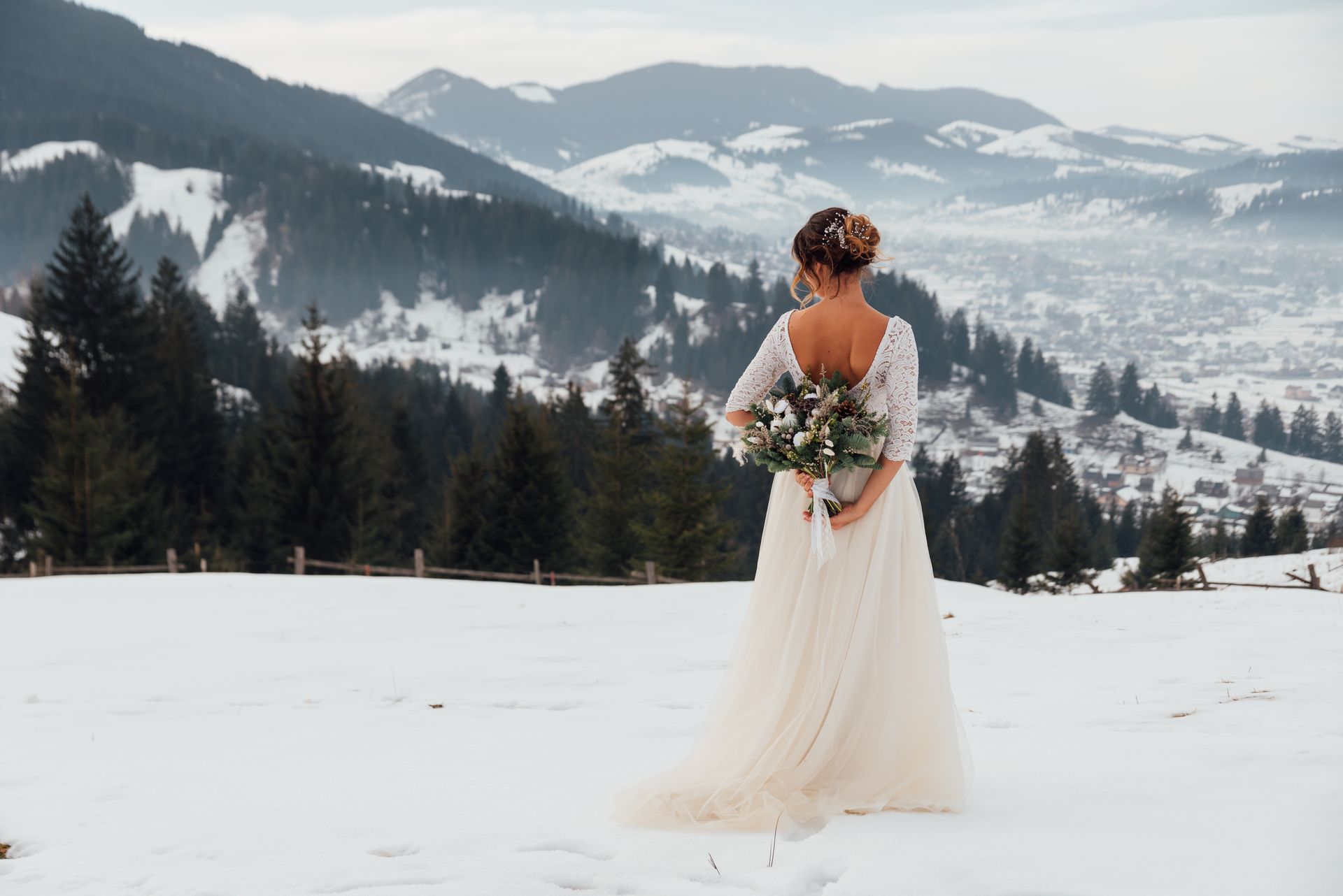 Bride in a white gown holding flowers, standing in a snowy mountain landscape