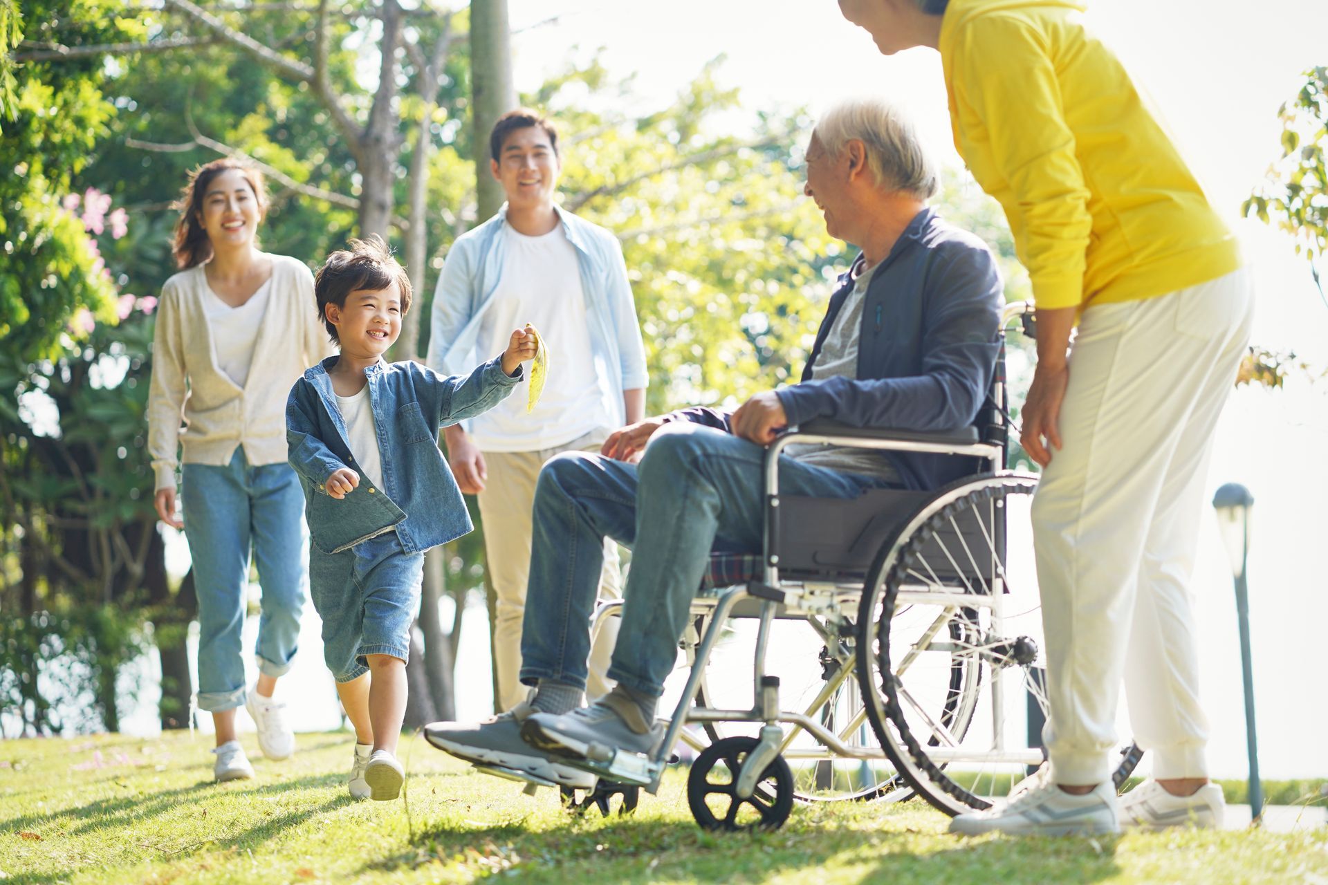Group walking with a man in a wheelchair in a sunny park, with one person pushing him.