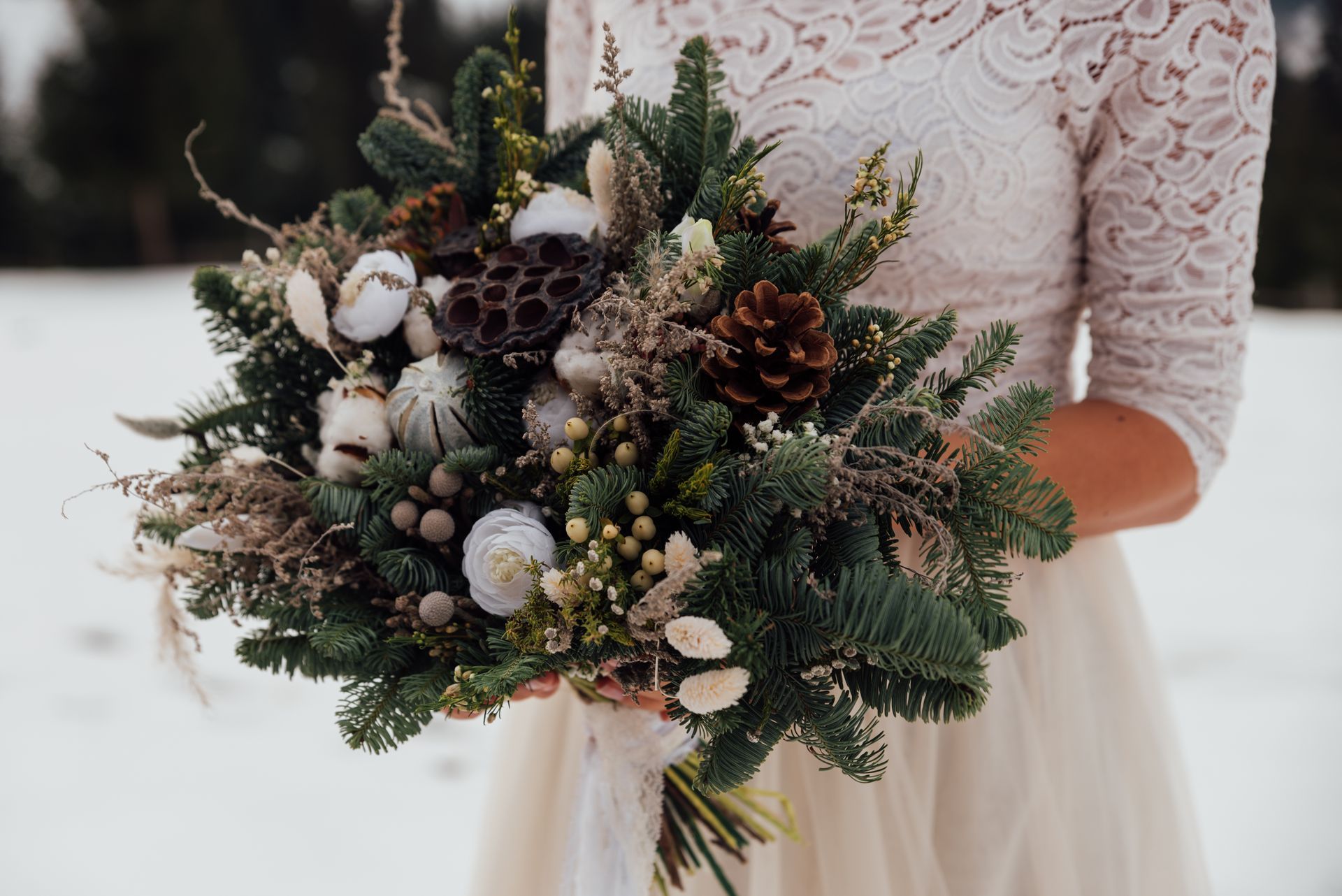 Bride holding a winter bouquet with pinecones, white ornaments, and greenery in a snowy setting