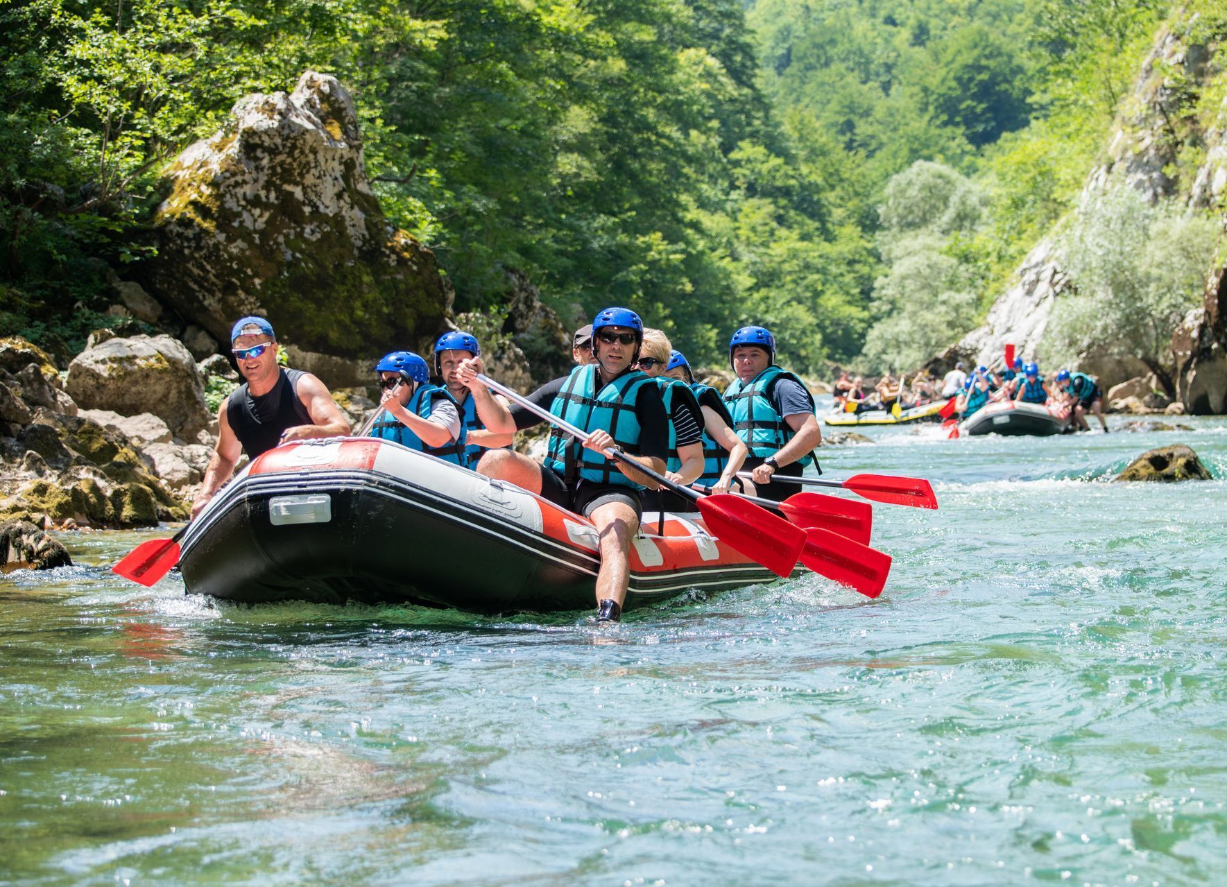 People rafting down a river. They are wearing life vests and using paddles. Green water and trees are visible.