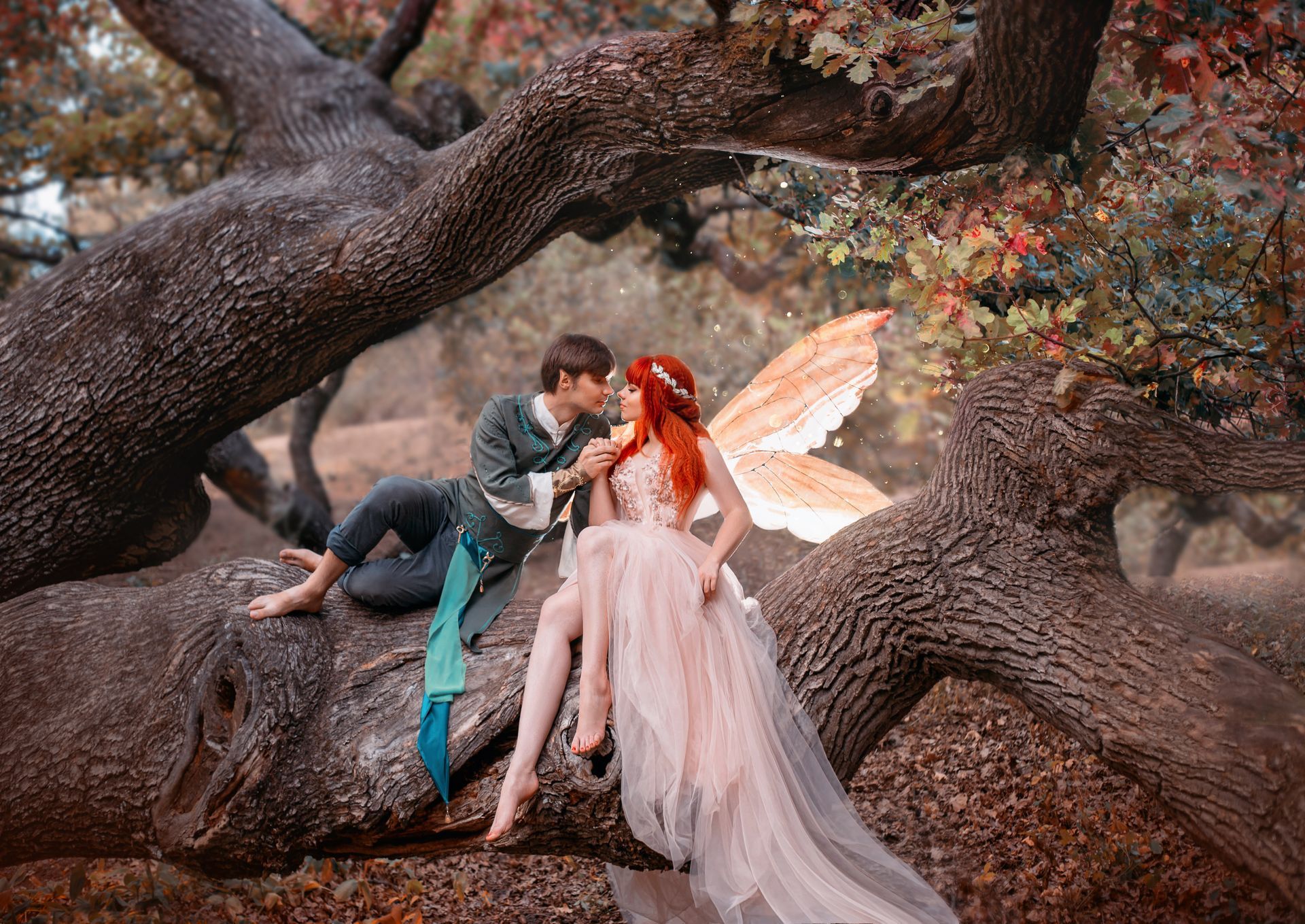 Couple in fantasy costumes sitting on tree branches in a wooded setting, with fairy wings and a pink gown
