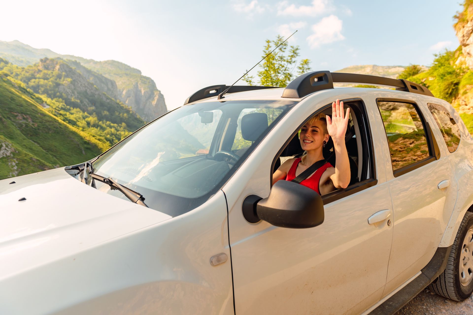 Woman waving from a white SUV, driving on a mountain road. Sunny day, green hills in the background.
