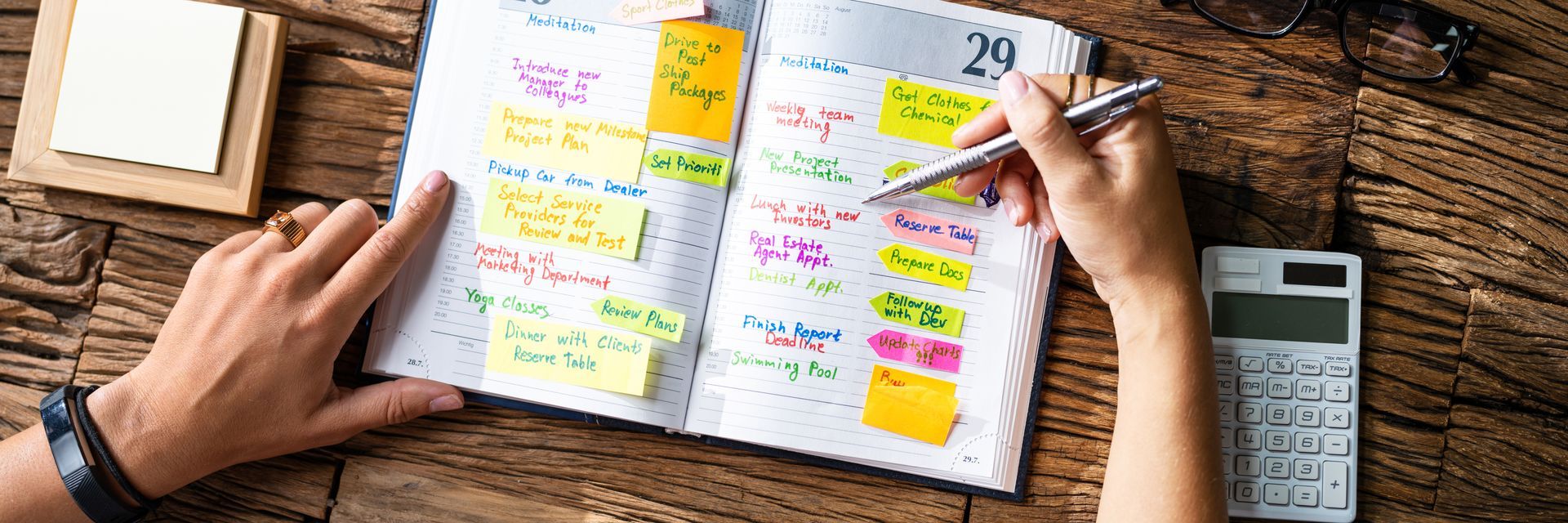 Hands planning in an open notebook with colorful sticky notes, pen, coffee, and calculator on a wooden desk