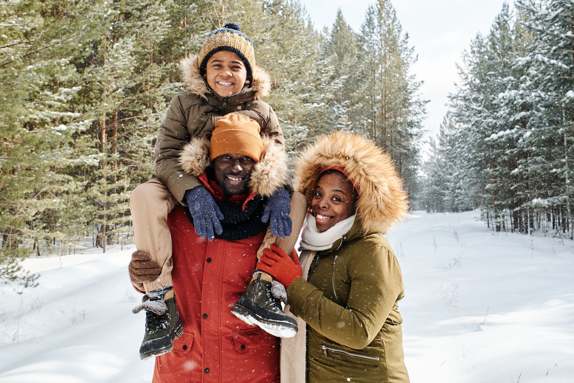 Family of three in winter coats smile in a snowy forest; child on father's shoulders.