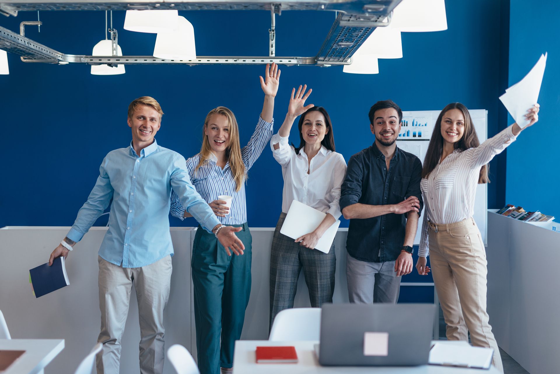 Group of coworkers standing in an office, smiling and raising their hands behind laptops and papers.