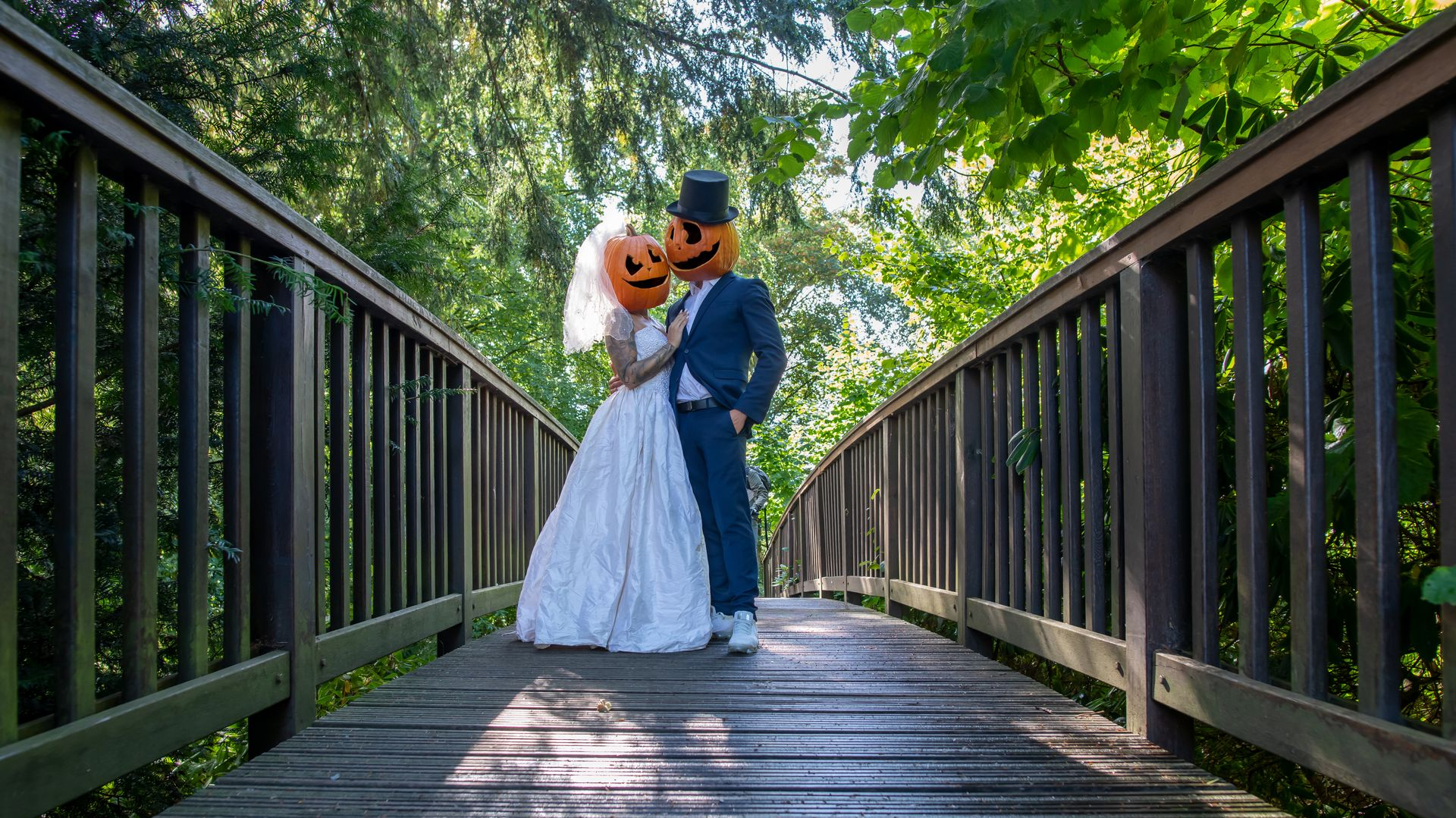 Bride and groom in wedding attire standing on a wooden bridge under green trees
