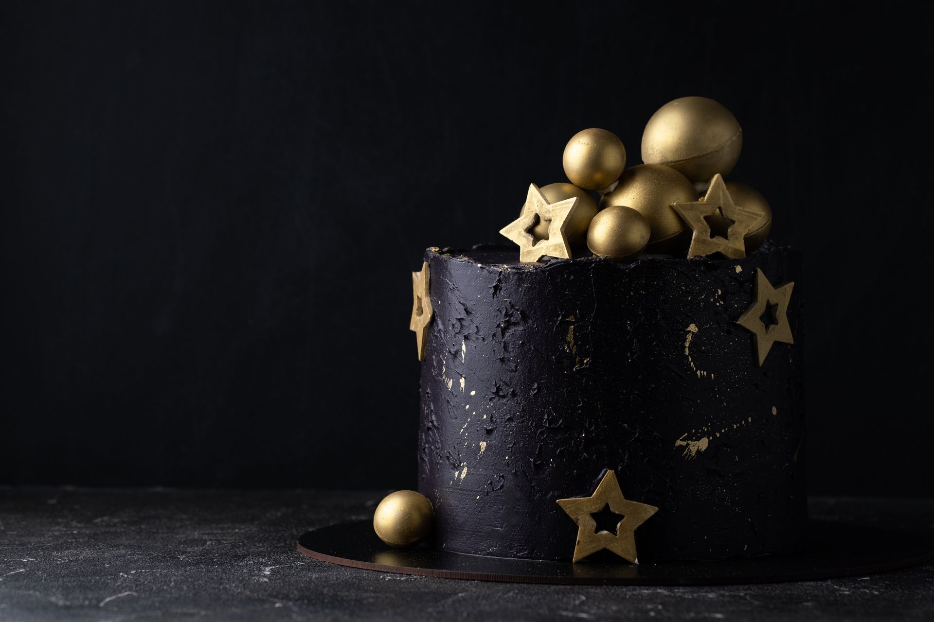 Black cake decorated with gold stars and metallic balls on a dark background