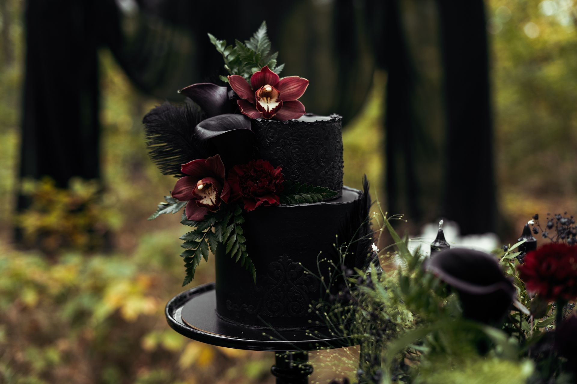 Black tiered cake with red flowers and greenery on a stand in a blurred outdoor setting
