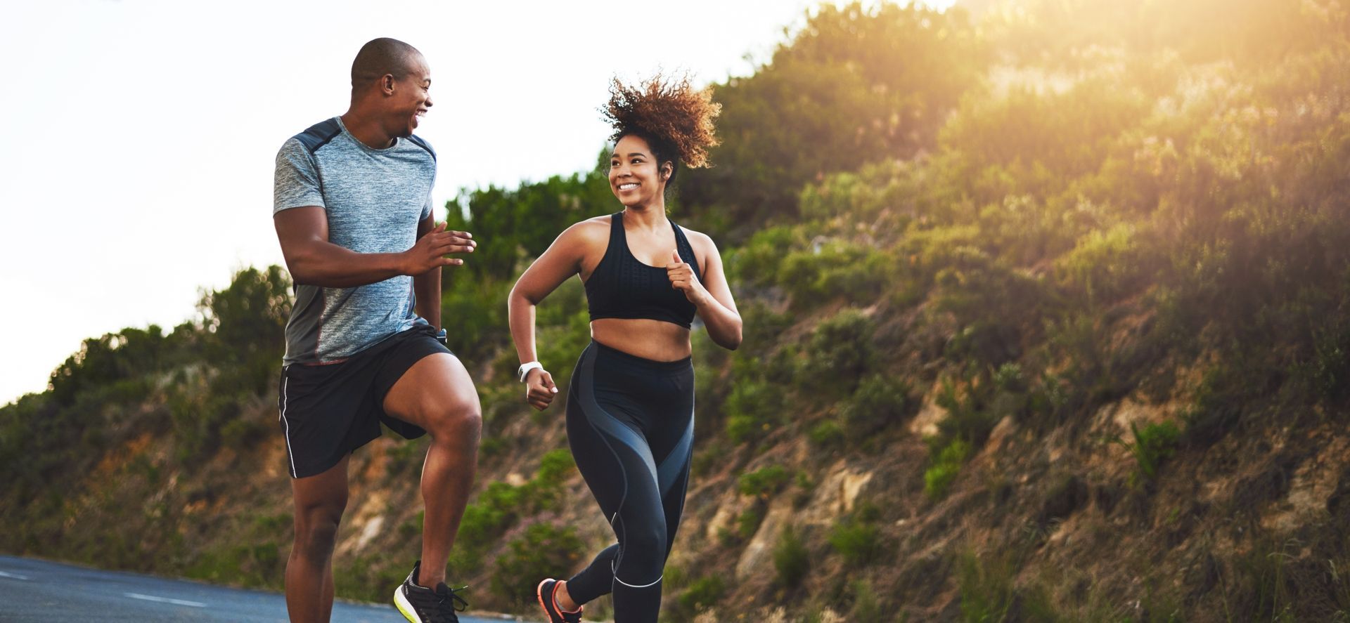 Two people running on a road, a woman in a sports bra smiles as a man runs beside her, sunlit hillside in the background.
