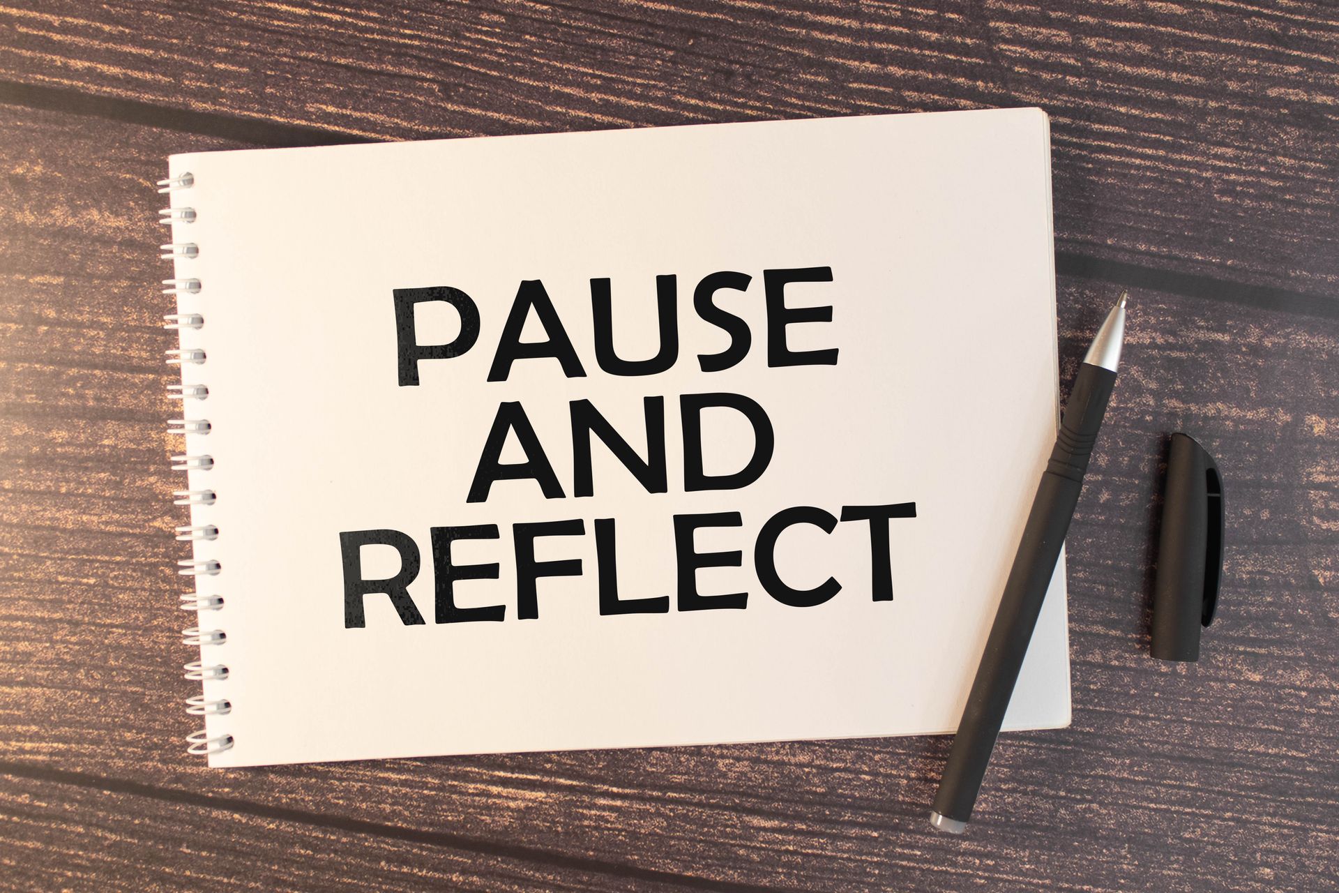 Notebook page with “PAUSE AND REFLECT” on a wooden desk beside a pen and marker.