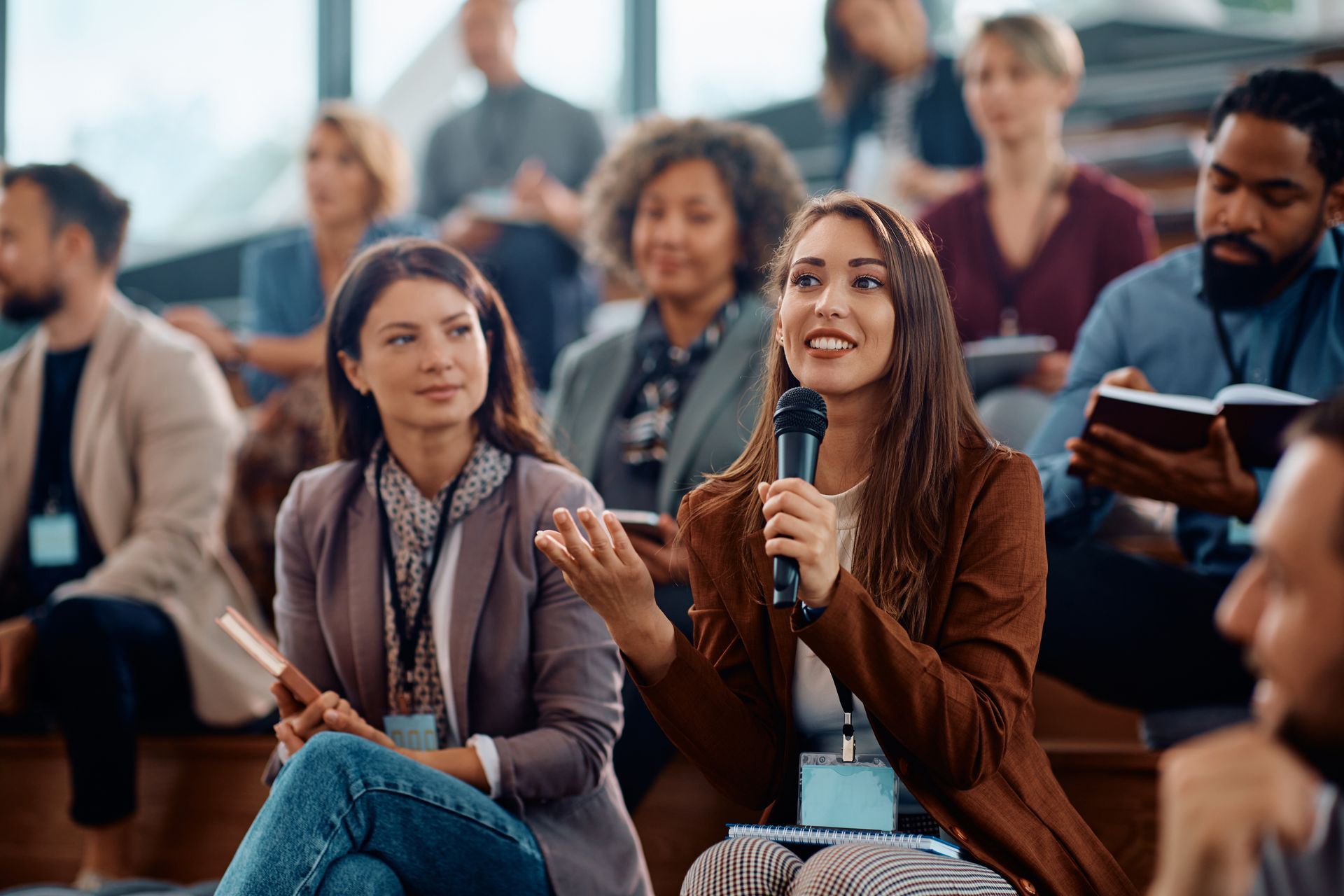 Woman speaking into a microphone at a seated audience discussion, with listeners in a bright indoor setting