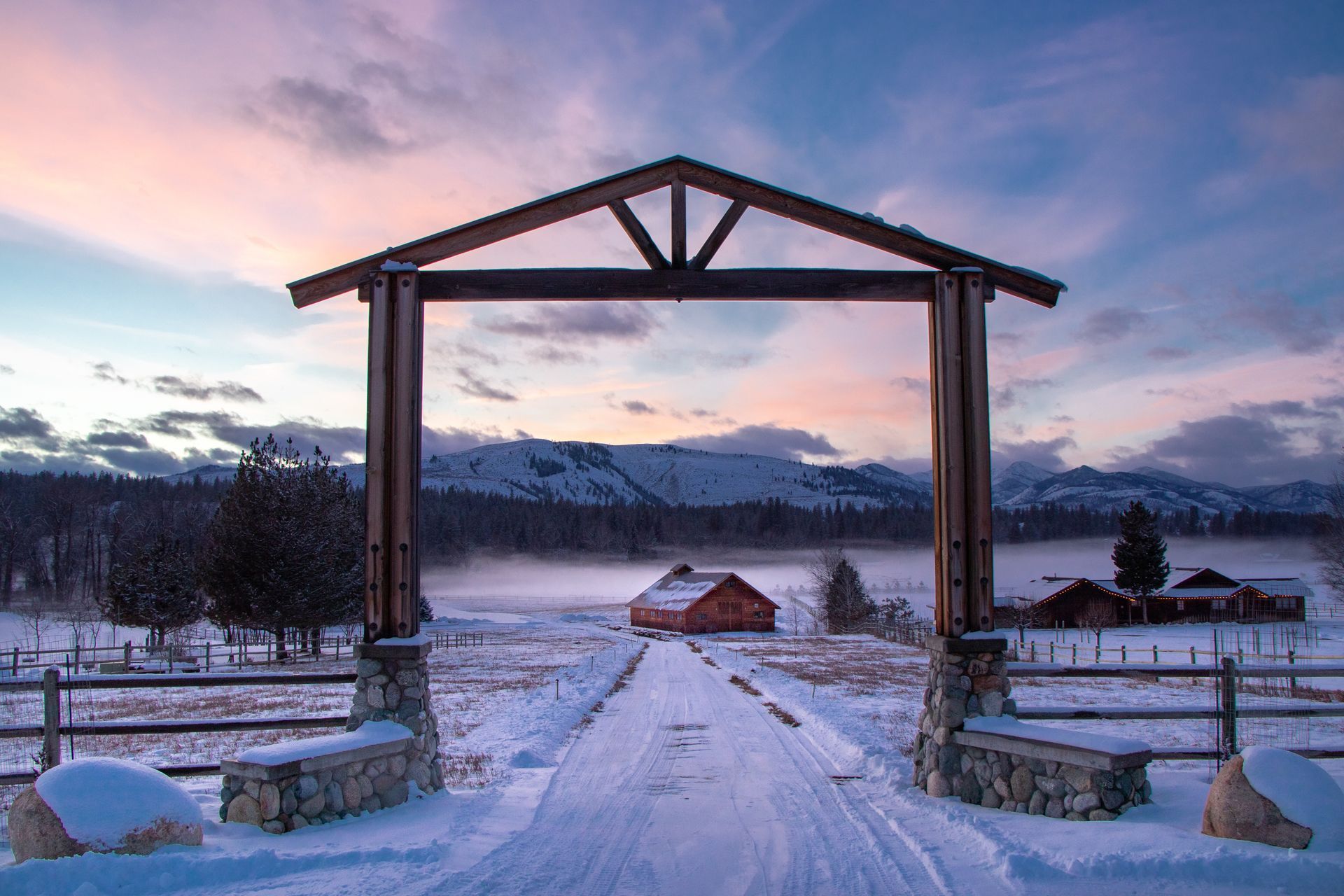 Snowy ranch entrance with a wooden archway and a view of mountains at sunrise.