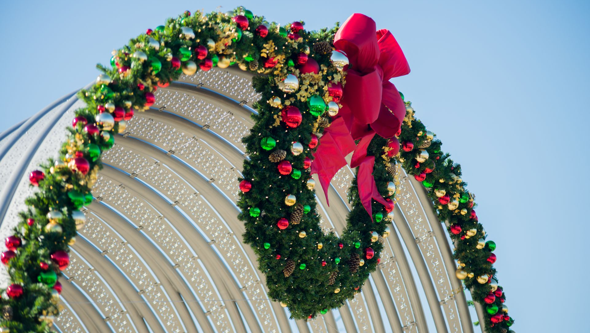 Christmas wreath on a white arched structure against a blue sky, decorated with red and green ornaments and a large red bow.