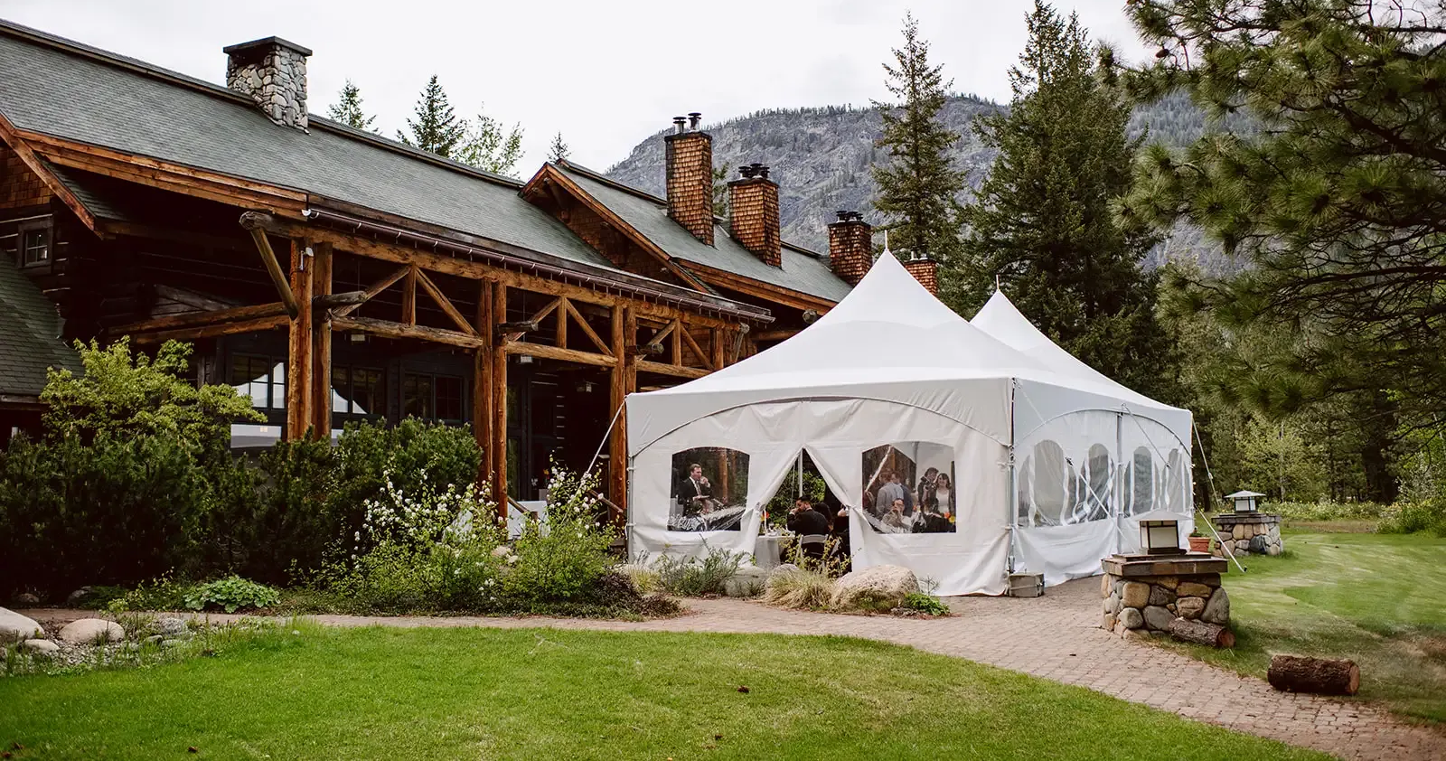 White event tent beside a rustic lodge with wooden porch, set on a grassy lawn near mountains