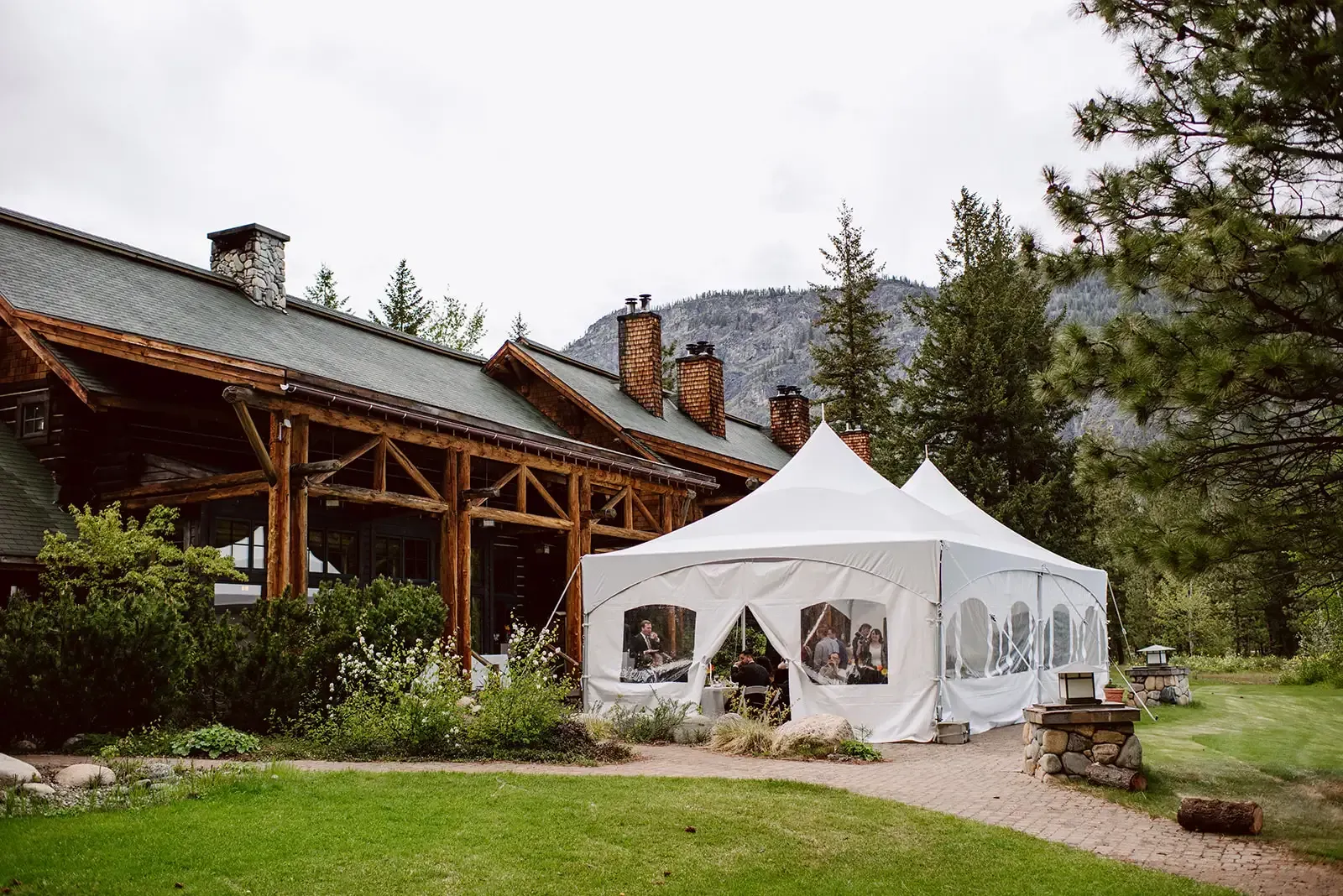 White event tent beside a rustic lodge on a grassy lawn, with mountains and trees in the background