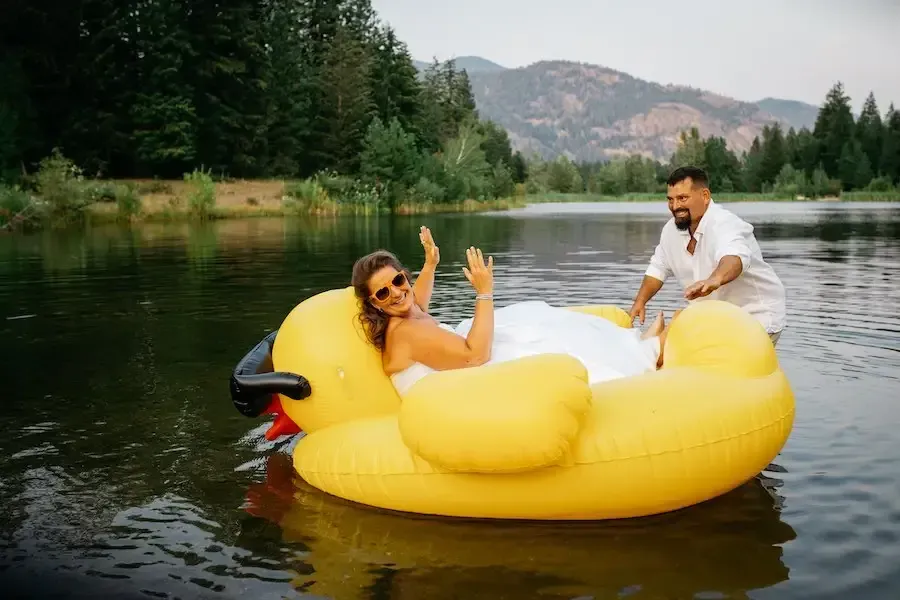 Bride in white dress waves from giant inflatable duck in lake, groom pushes. Mountains and trees in background.