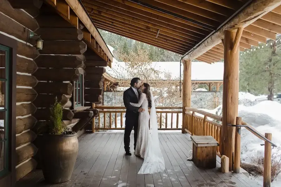 Couple in wedding attire kissing on a snowy cabin porch with wooden railings and rustic beams