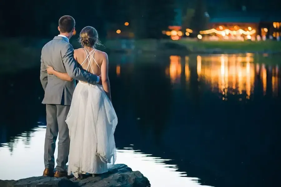A bride and groom are standing on a rock overlooking a lake at night.
