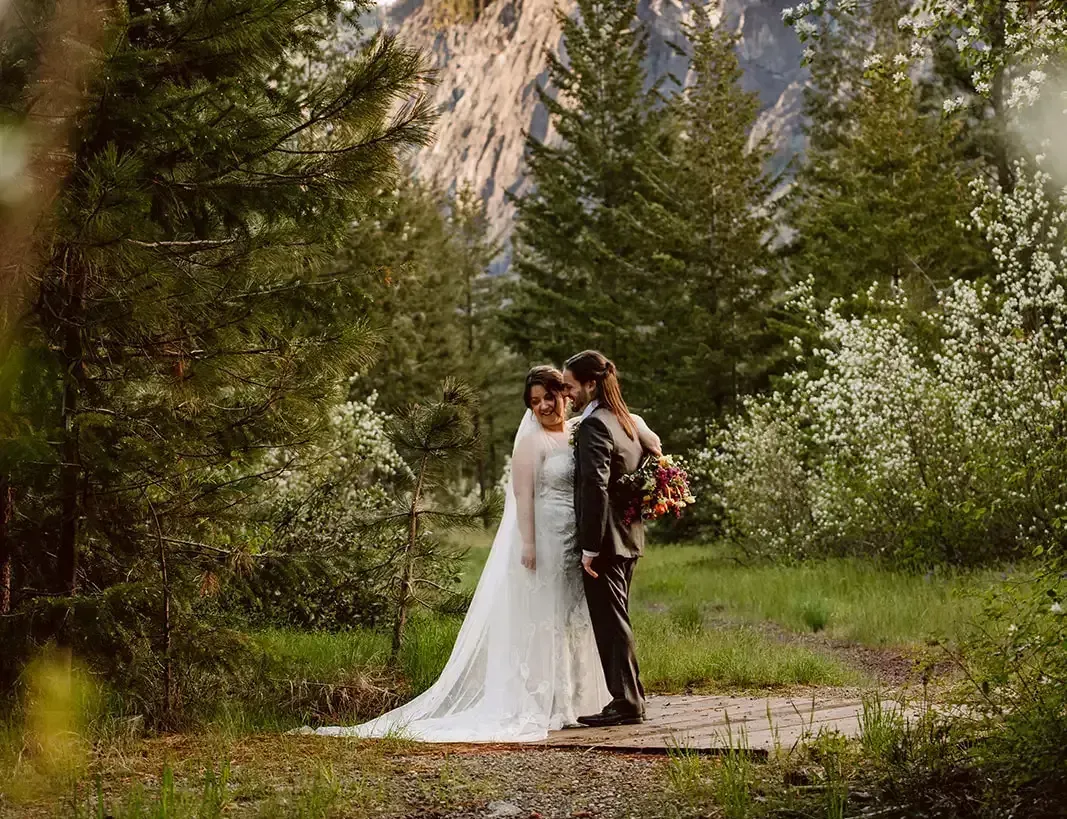 A bride and groom are standing in the woods with a mountain in the background.