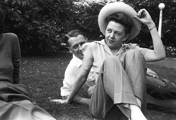 Man and woman seated on grass, woman adjusting a hat, man smiling.