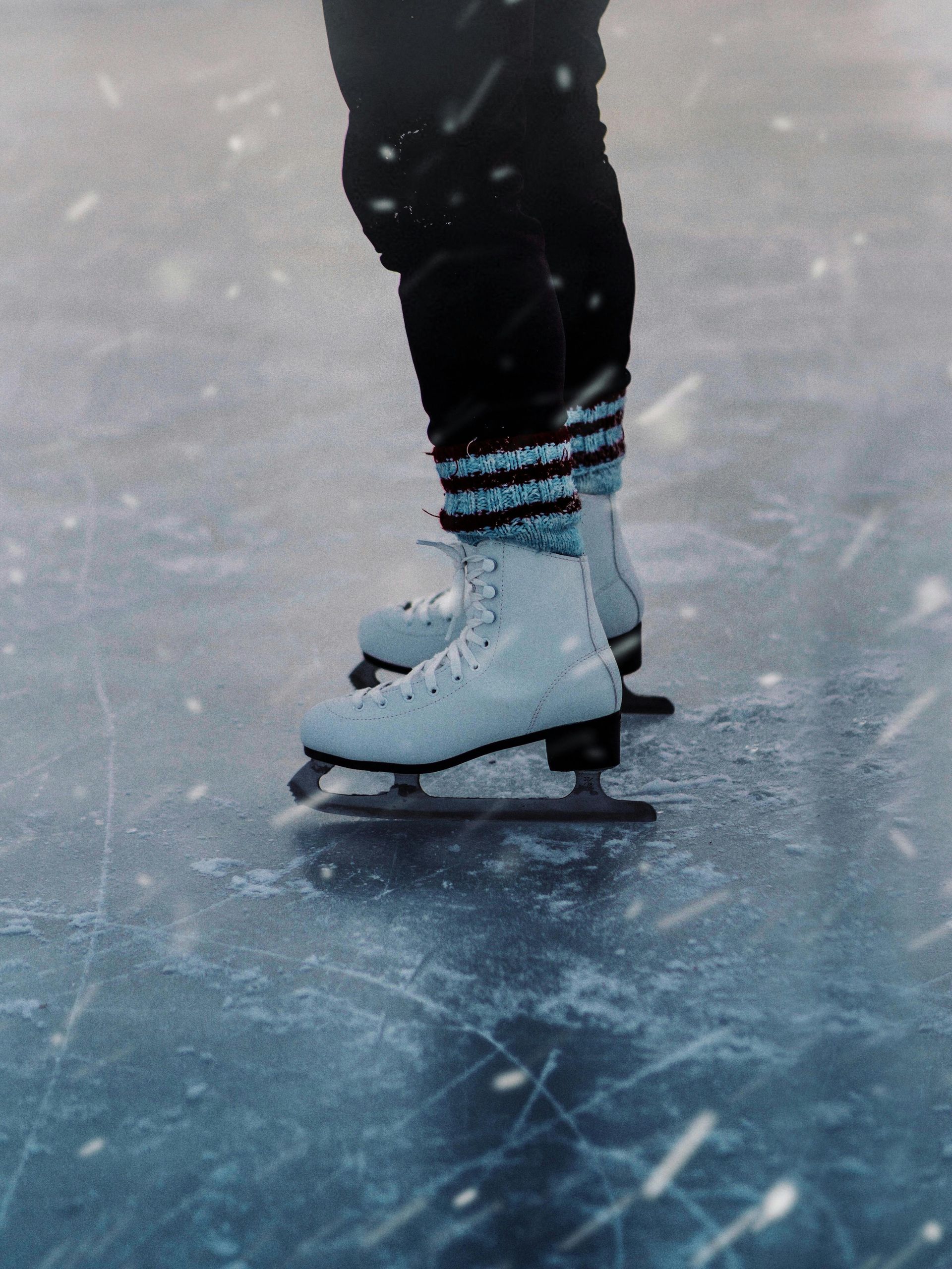 Ice skater's feet on ice. White skates, dark pants with blue striped leg warmers.