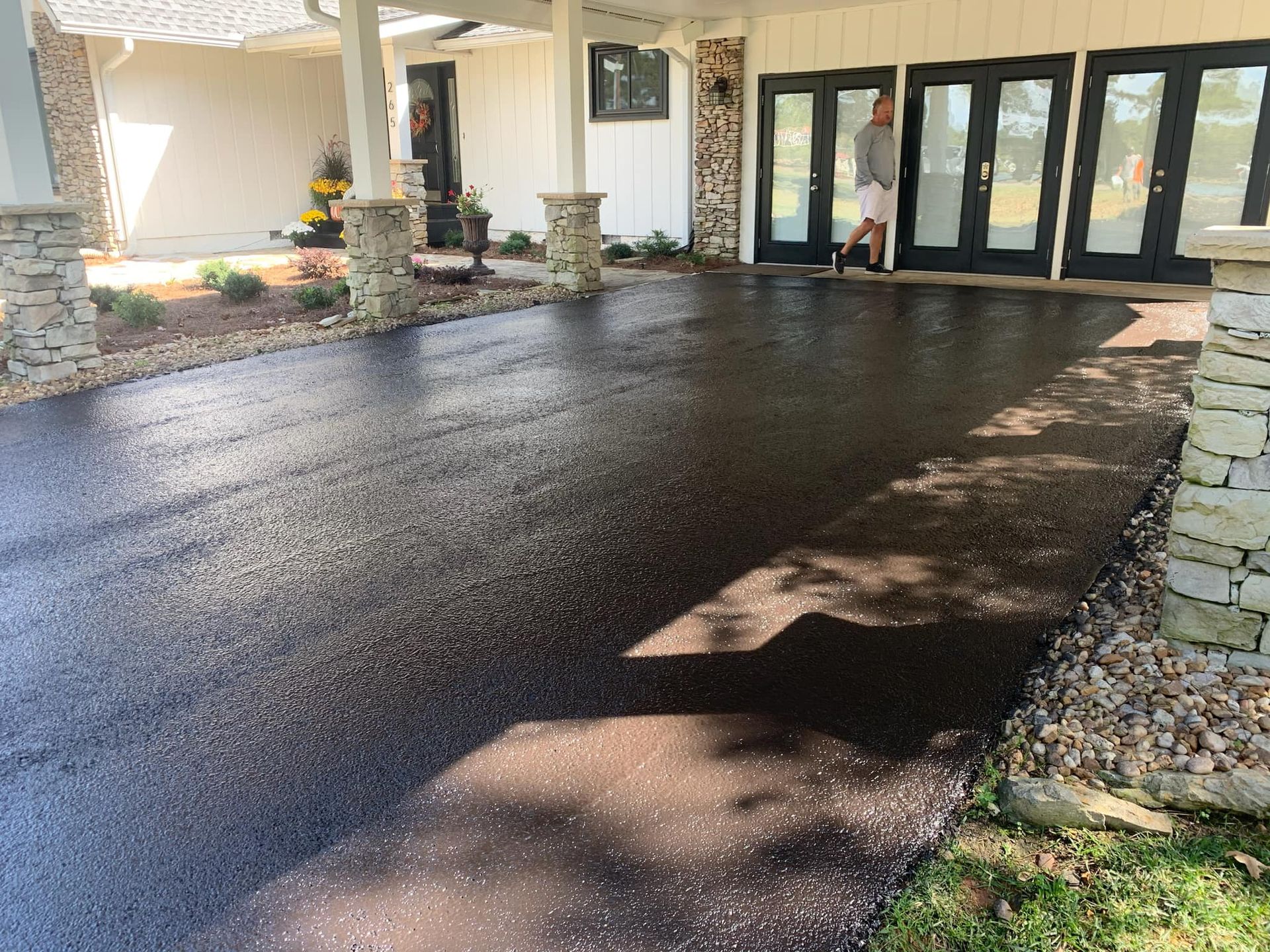 A newly paved, dark asphalt driveway under a stone-pillared portico leading to a building entrance with glass doors.